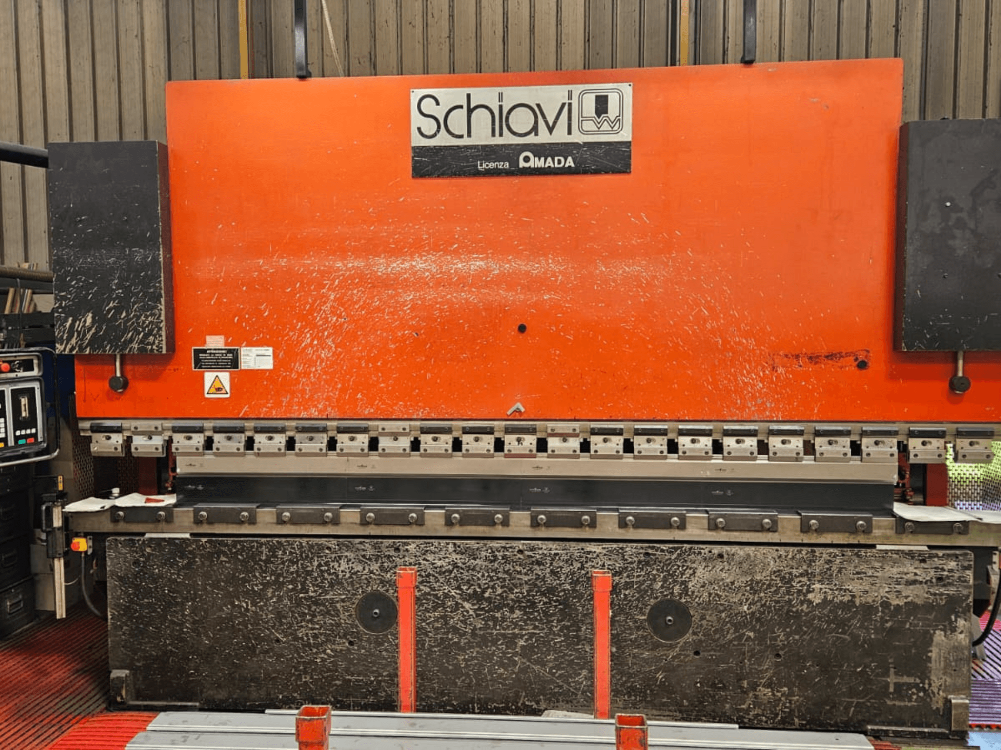 Máquina curvadora de metales Schiavi Amada, vista frontal, con cuerpo naranja, panel de control a la izquierda y múltiples herramientas de curvado visibles.