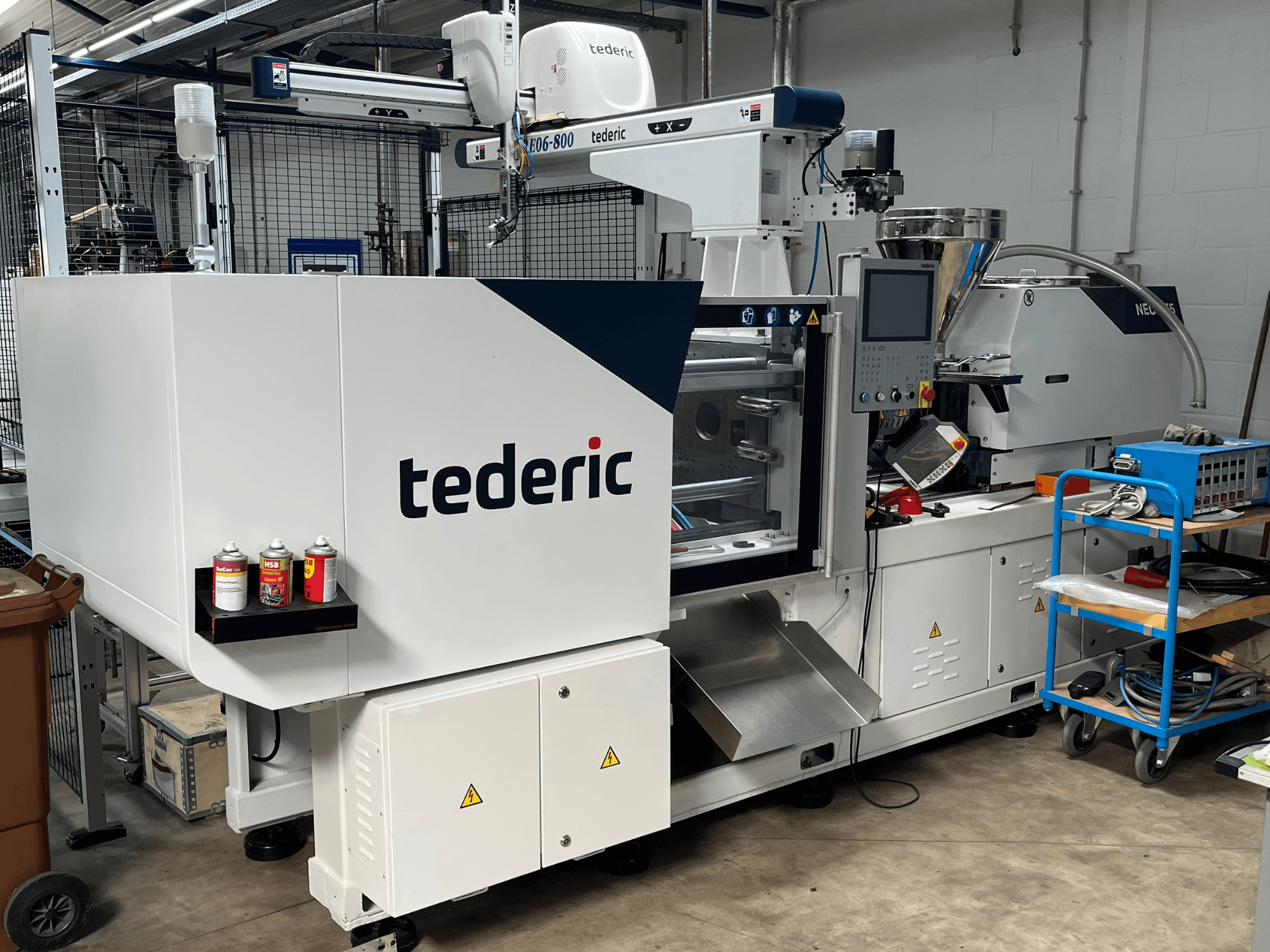 Máquina de moldeo por inyección hidráulica TEDERIC NEO.E55/e110h en un taller, vista lateral con el panel de control y las herramientas adyacentes.
