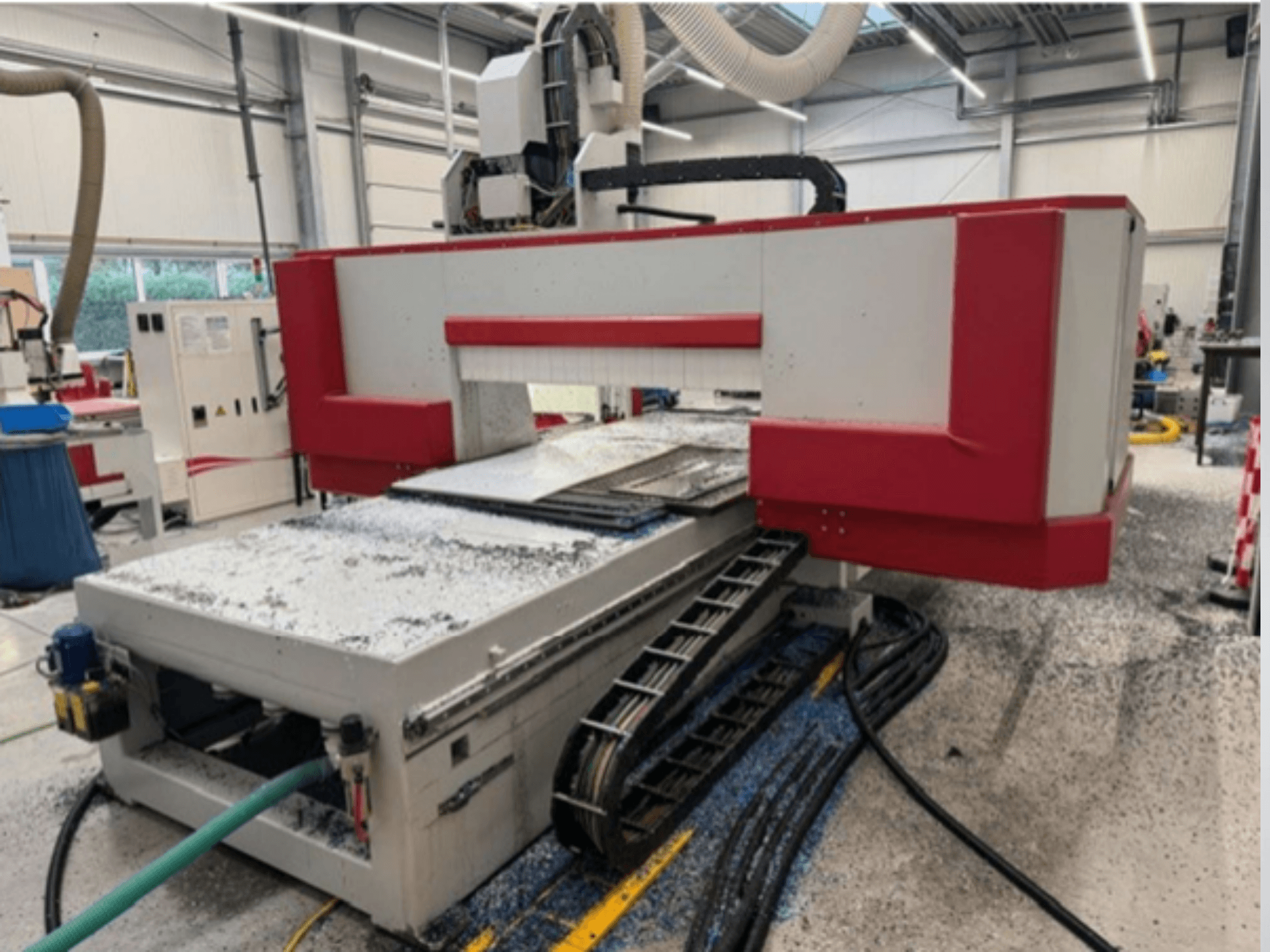 Máquina CNC roja y gris en un taller, vista lateral mostrando la mesa de material, la manguera de refrigerante y el serrín en el suelo.