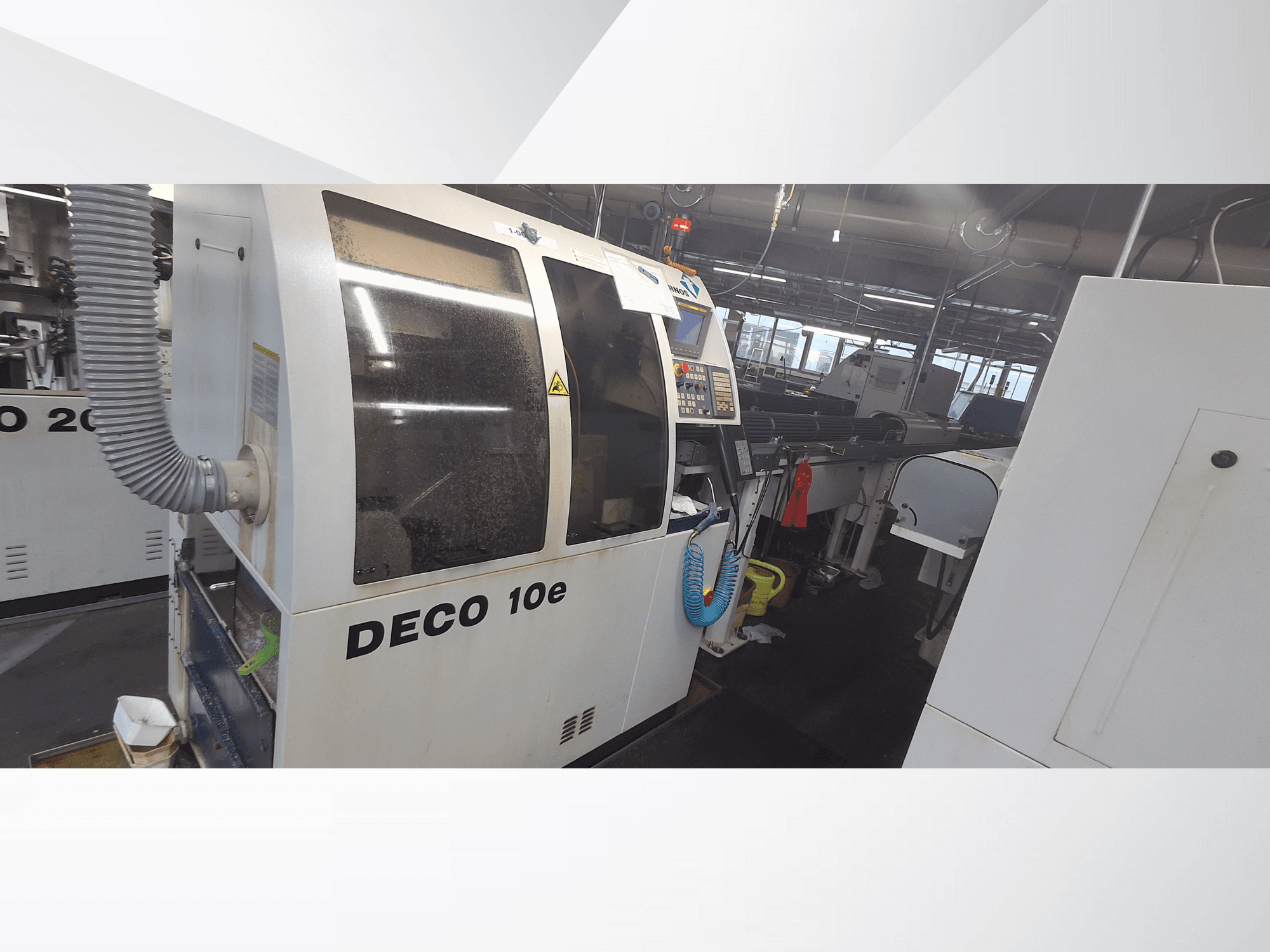 Máquina DECO 10e en un taller; vista lateral que muestra el panel de control y la manguera de ventilación, con maquinaria borrosa al fondo.