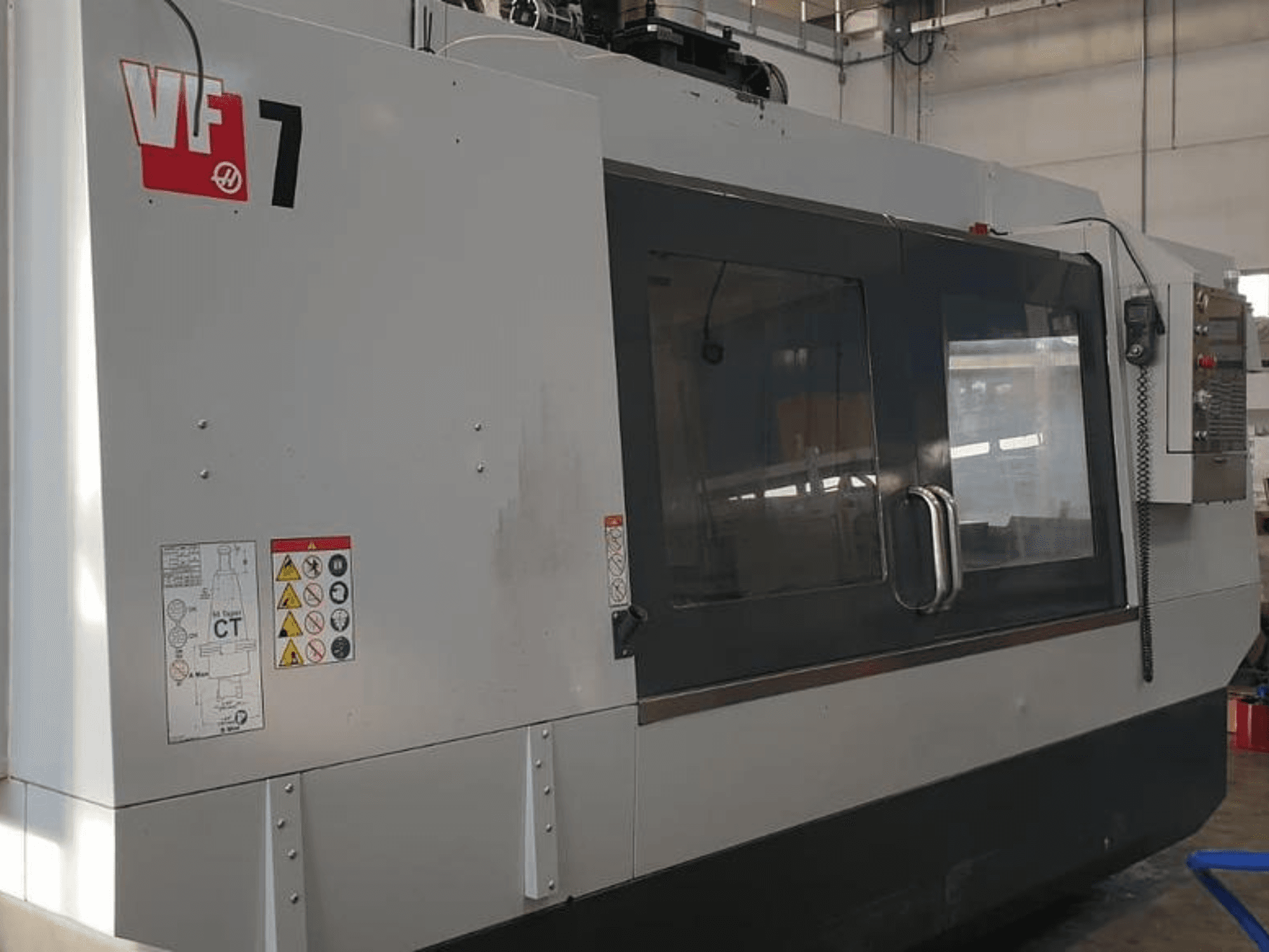 Máquina CNC Haas VF-7 en vista lateral, con acabado gris metalizado, panel de control y etiquetas de seguridad.