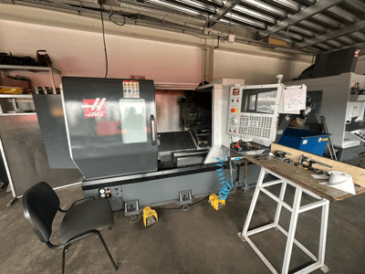 Torno CNC Haas (modelo ST-10) en un taller, vista lateral mostrando controles, herramientas en un banco de trabajo y una silla cerca.