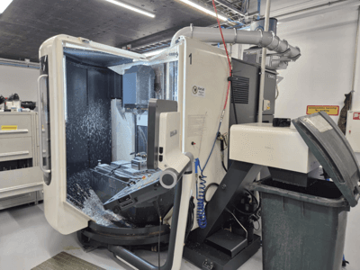 Fresadora DMG abierta con pulverización de refrigerante, mostrando las virutas de metal y los controles de la herramienta, vista de frente.