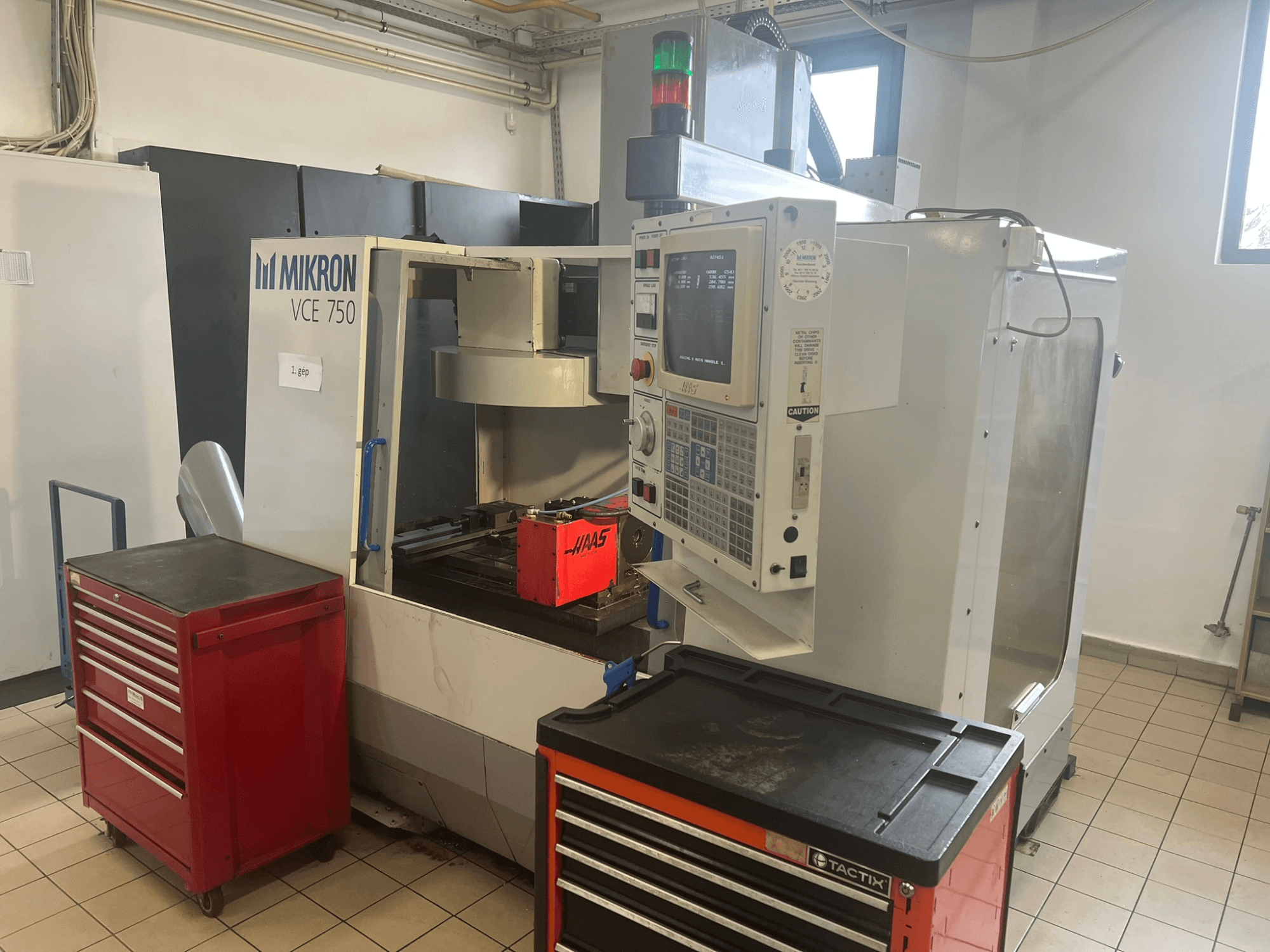 Alt text: "Centro de mecanizado vertical MIKRON VCE 750, vista frontal con panel de control y cajas de herramientas rojas, en un amplio taller".