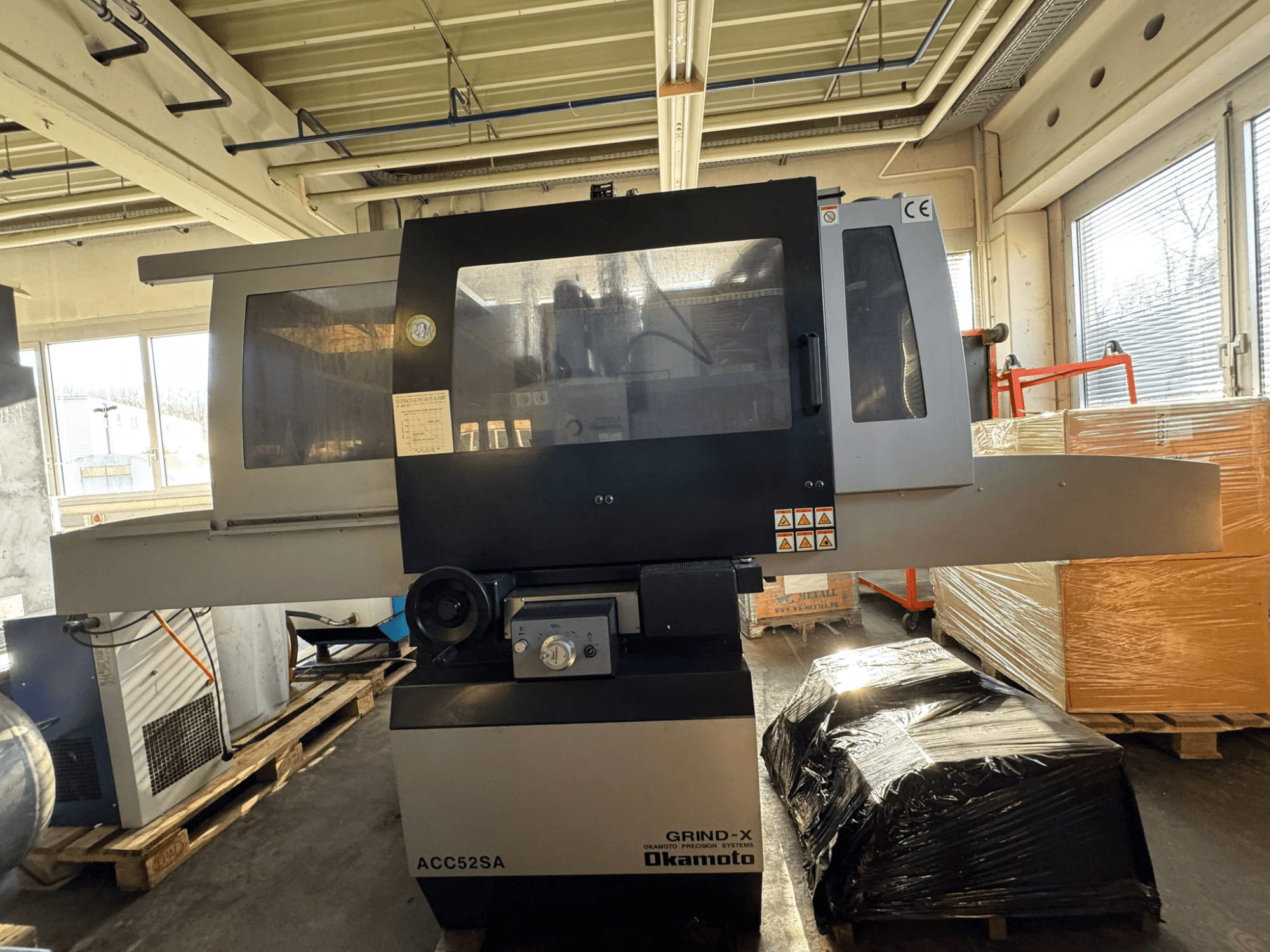 Máquina rectificadora plana Okamoto ACC 52 SA vista de perfil, en la que se aprecian los mandos y las etiquetas, rodeada de palés y cajas.
