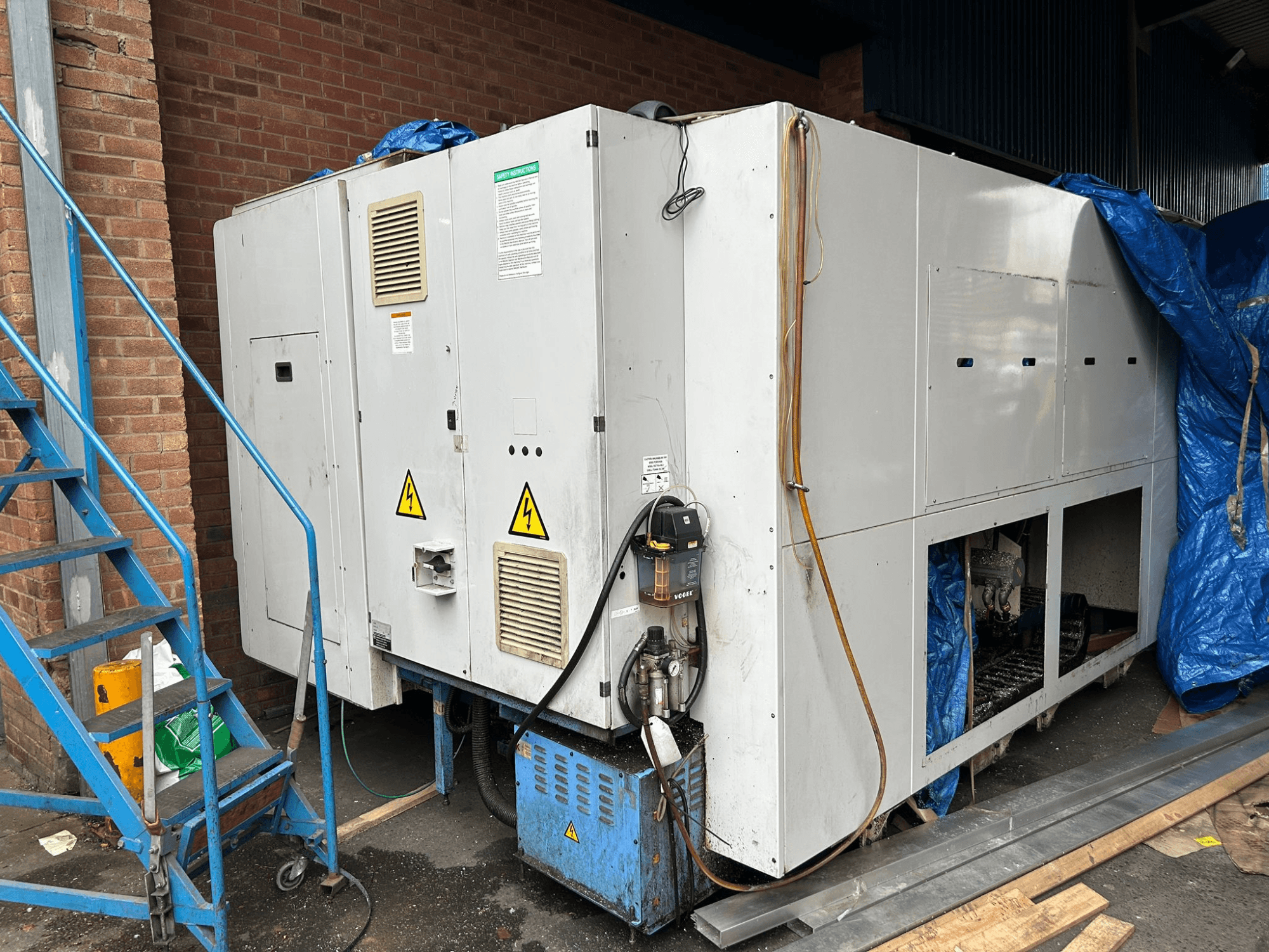 Vista lateral de una gran enfriadora industrial, marca y modelo poco claros, mostrando las conexiones eléctricas, las señales de advertencia y la cubierta de lona azul.