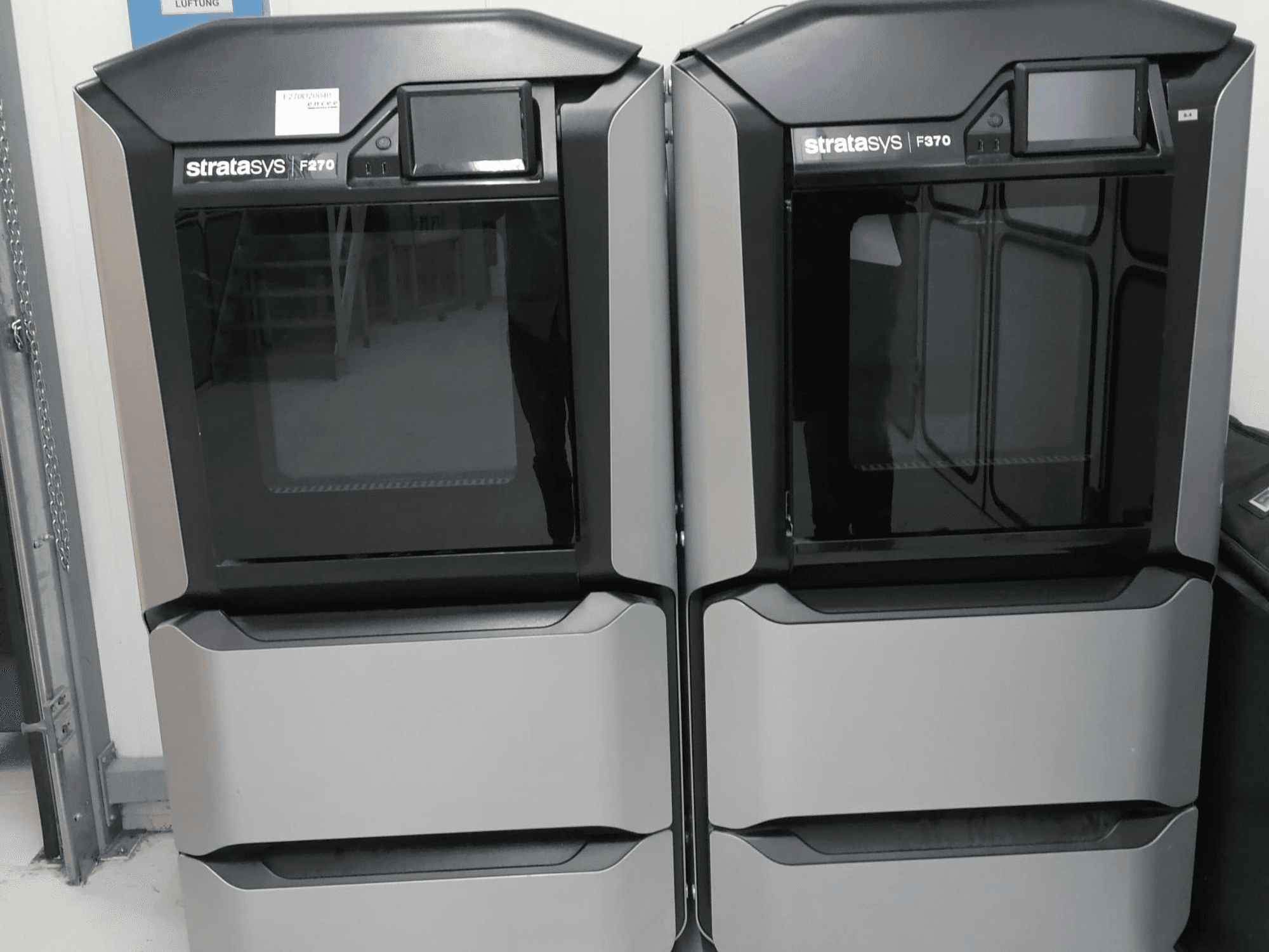 Vista frontal de la impresora 3D de plástico Stratasys F270, que muestra máquinas dobles con elegantes exteriores grises y pantallas digitales.