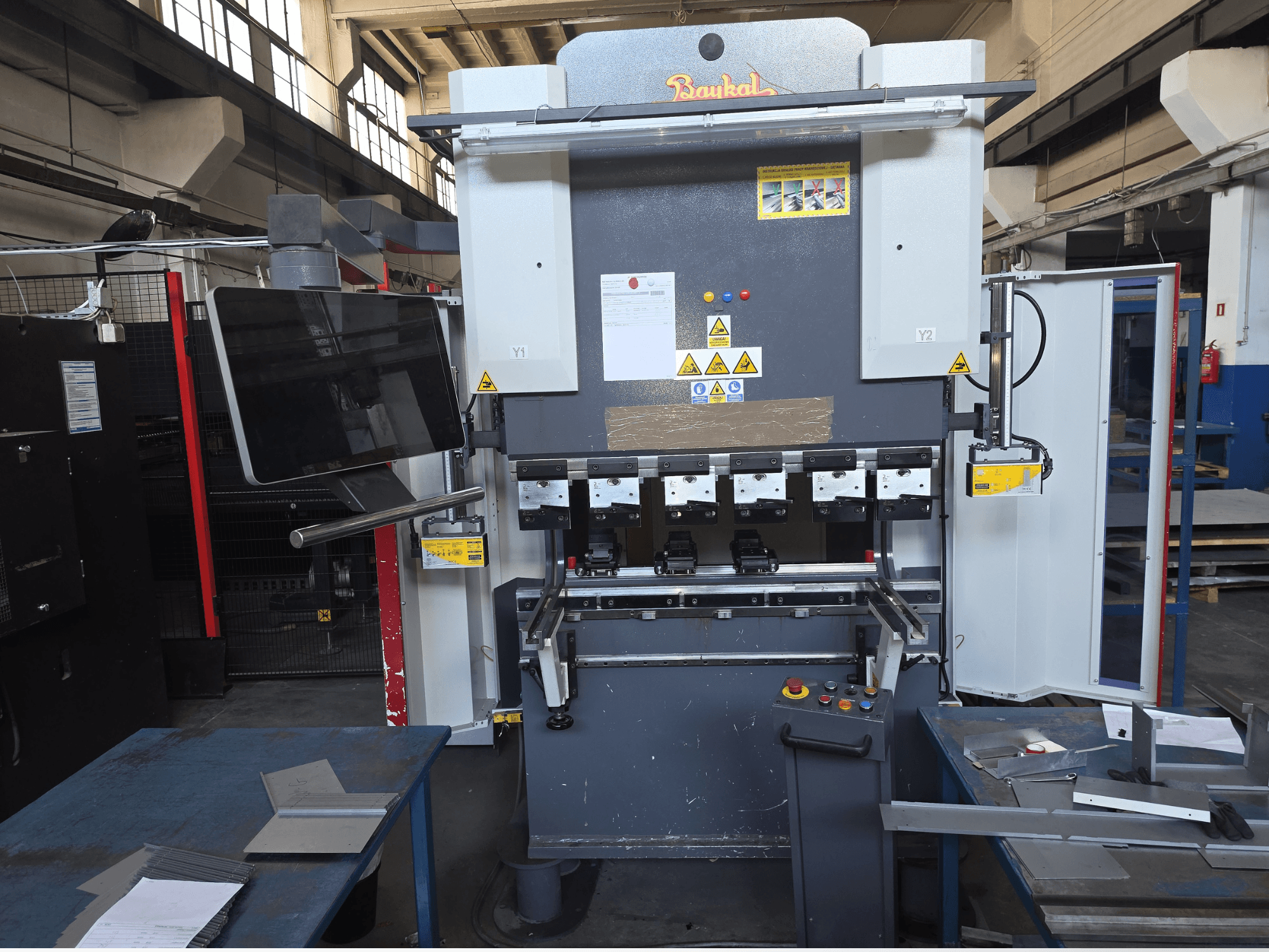 Plegadora Baykal APHS 125040, vista frontal, en la que se aprecian el panel de control y los dispositivos de seguridad, junto con los materiales sobre una mesa de trabajo azul.