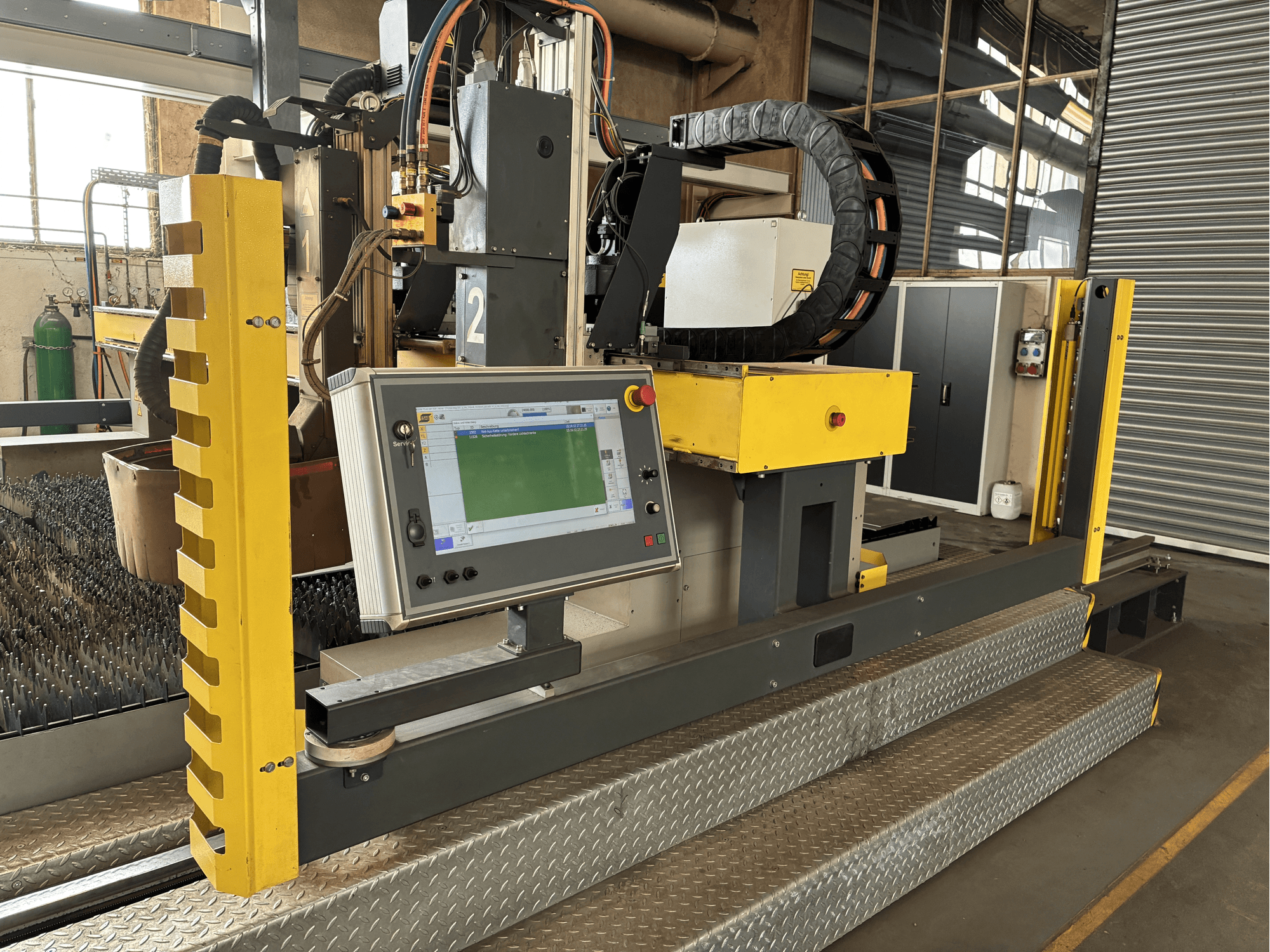 Máquina de corte industrial con panel de control táctil, protección de seguridad amarilla, vista frontal, base de rejilla metálica visible.