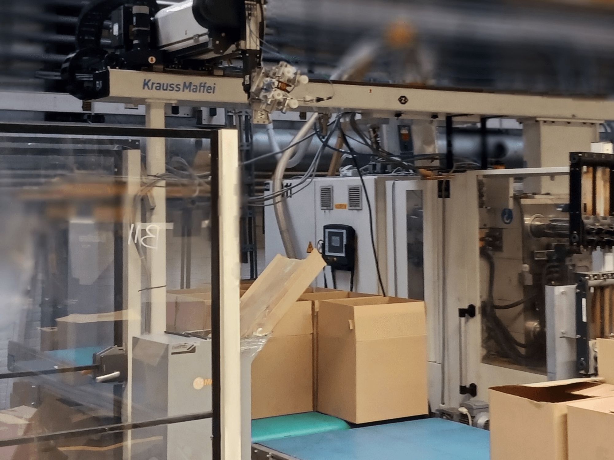 Sistema de automatización Krauss Maffei en una fábrica, vista frontal que muestra cajas en una cinta transportadora y componentes de maquinaria en funcionamiento.