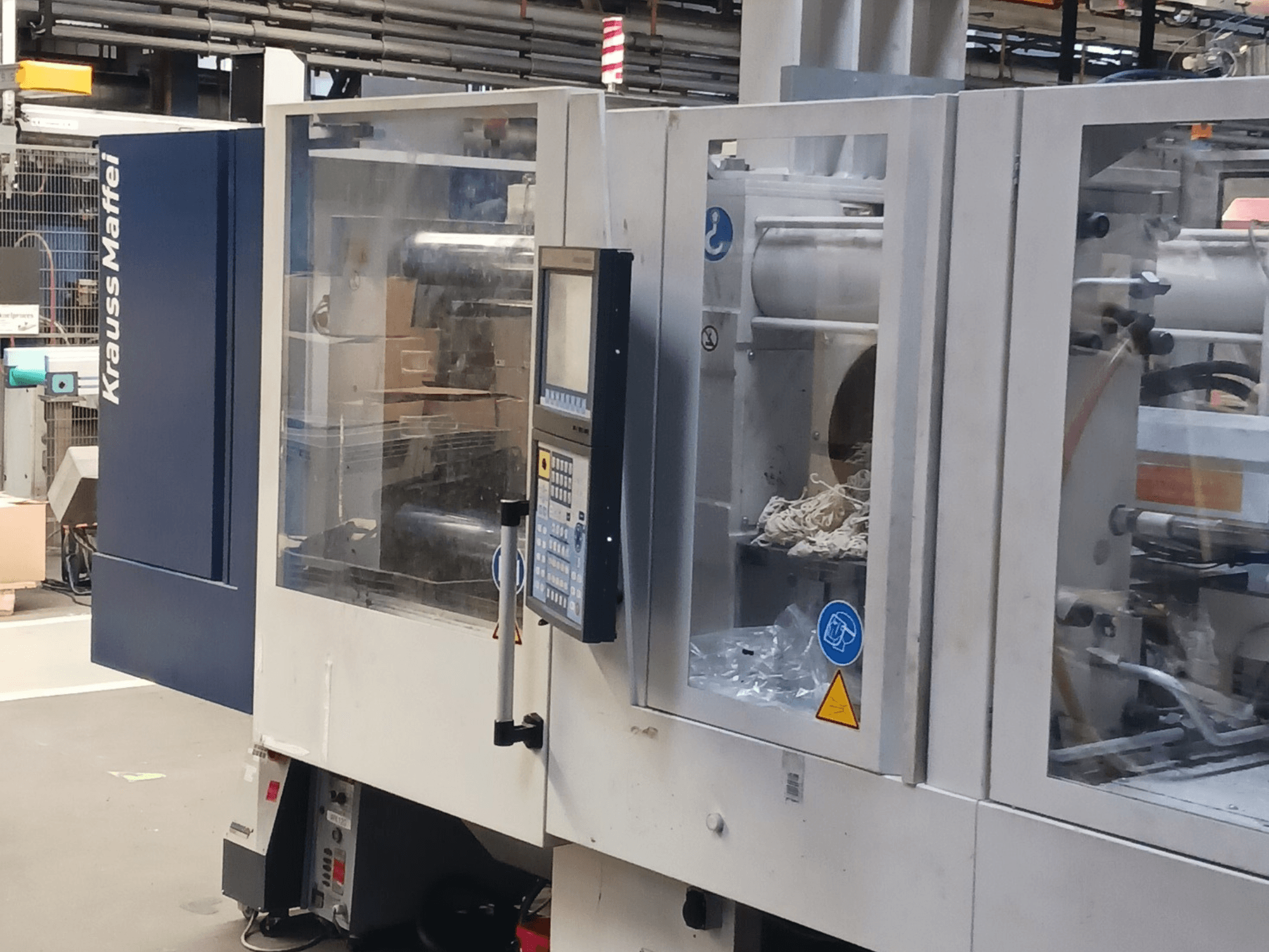 Máquina de moldeo por inyección KraussMaffei, vista lateral, mostrando el panel de control, la maquinaria y los materiales de embalaje al fondo.