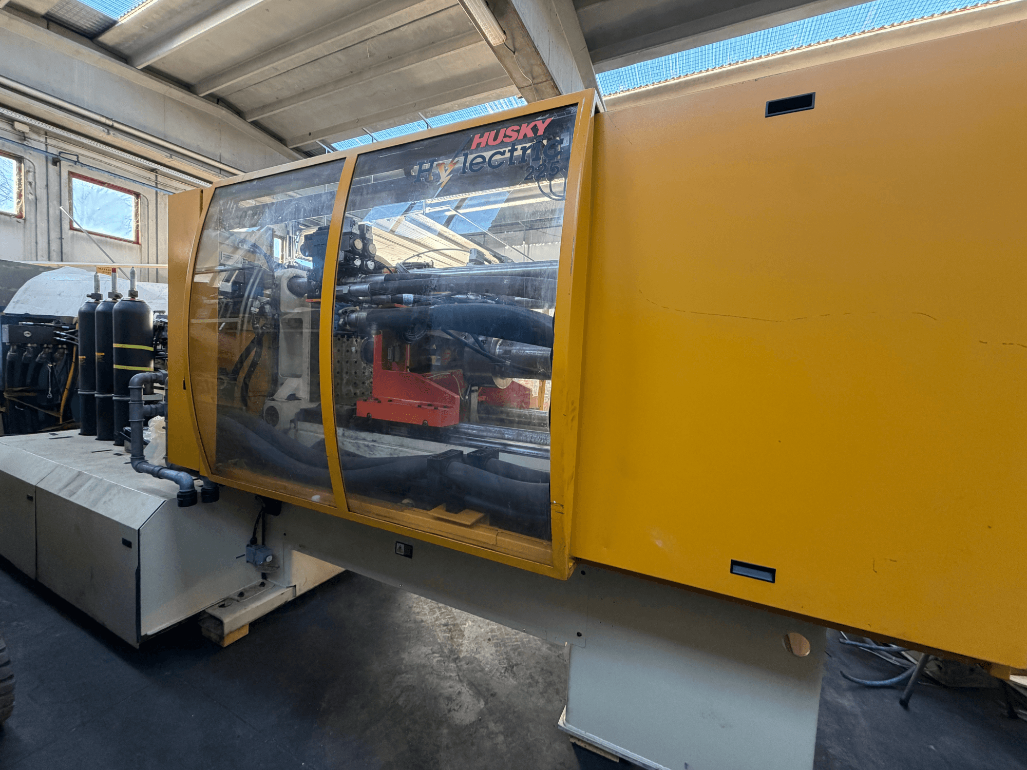 Máquina de moldeo por inyección eléctrica Husky HL225 RS65, vista frontal con carcasa amarilla, panel de control visible y componentes hidráulicos.