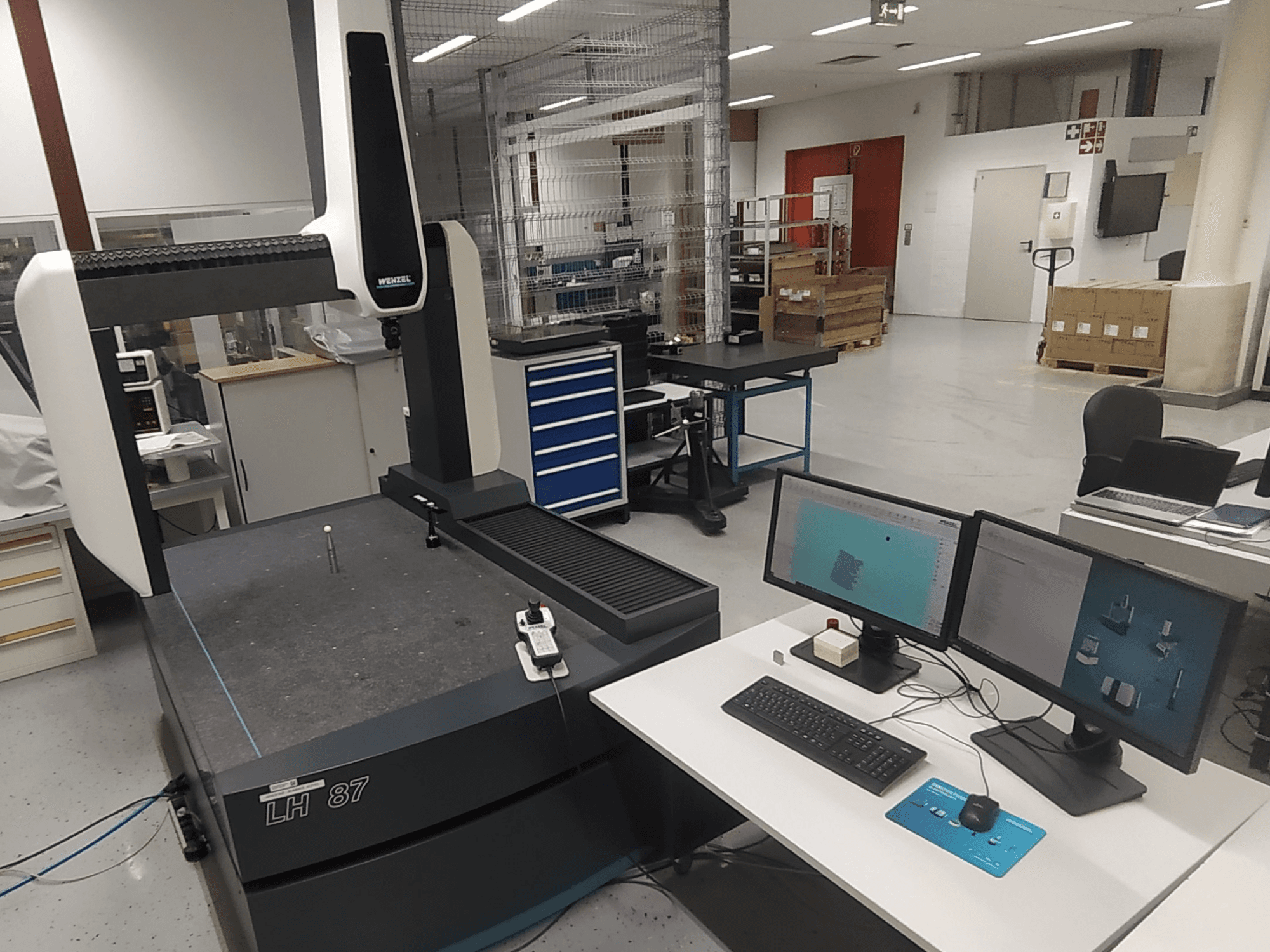 Máquina de medición por coordenadas Wenzel LH 87 visible en un laboratorio, con monitores dobles y sonda de medición sobre la mesa.
