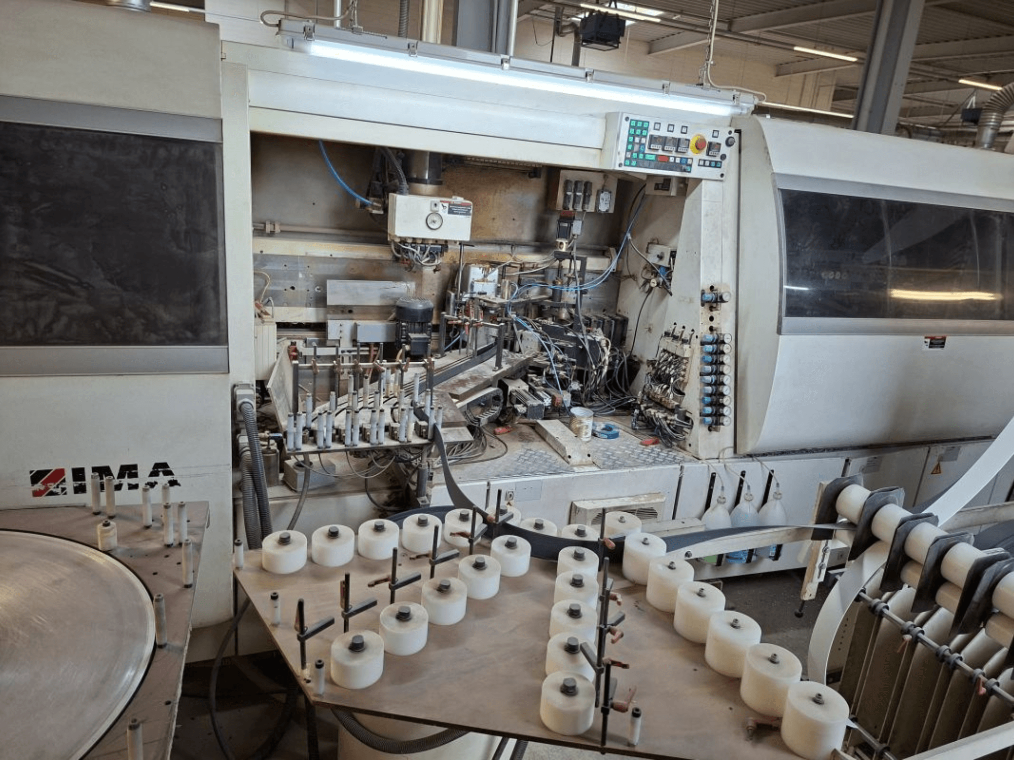 Maquinaria IMA vista desde un lateral, mostrando complejos componentes mecánicos y una mesa redonda con bobinas blancas.