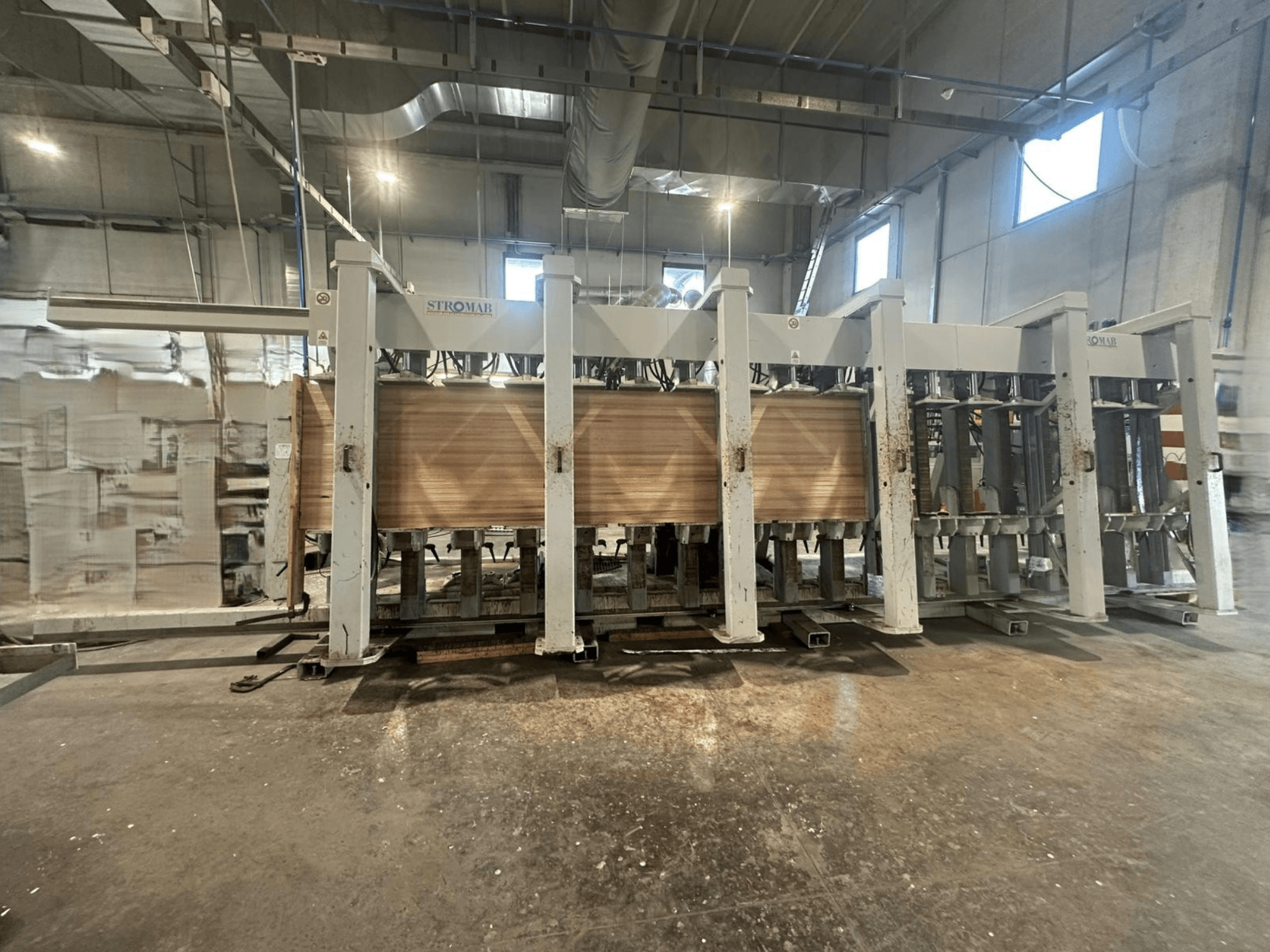 Máquina industrial STROMAB en un almacén, vista frontal, con paneles de madera en proceso, mostrando la estructura metálica y las piezas mecánicas.