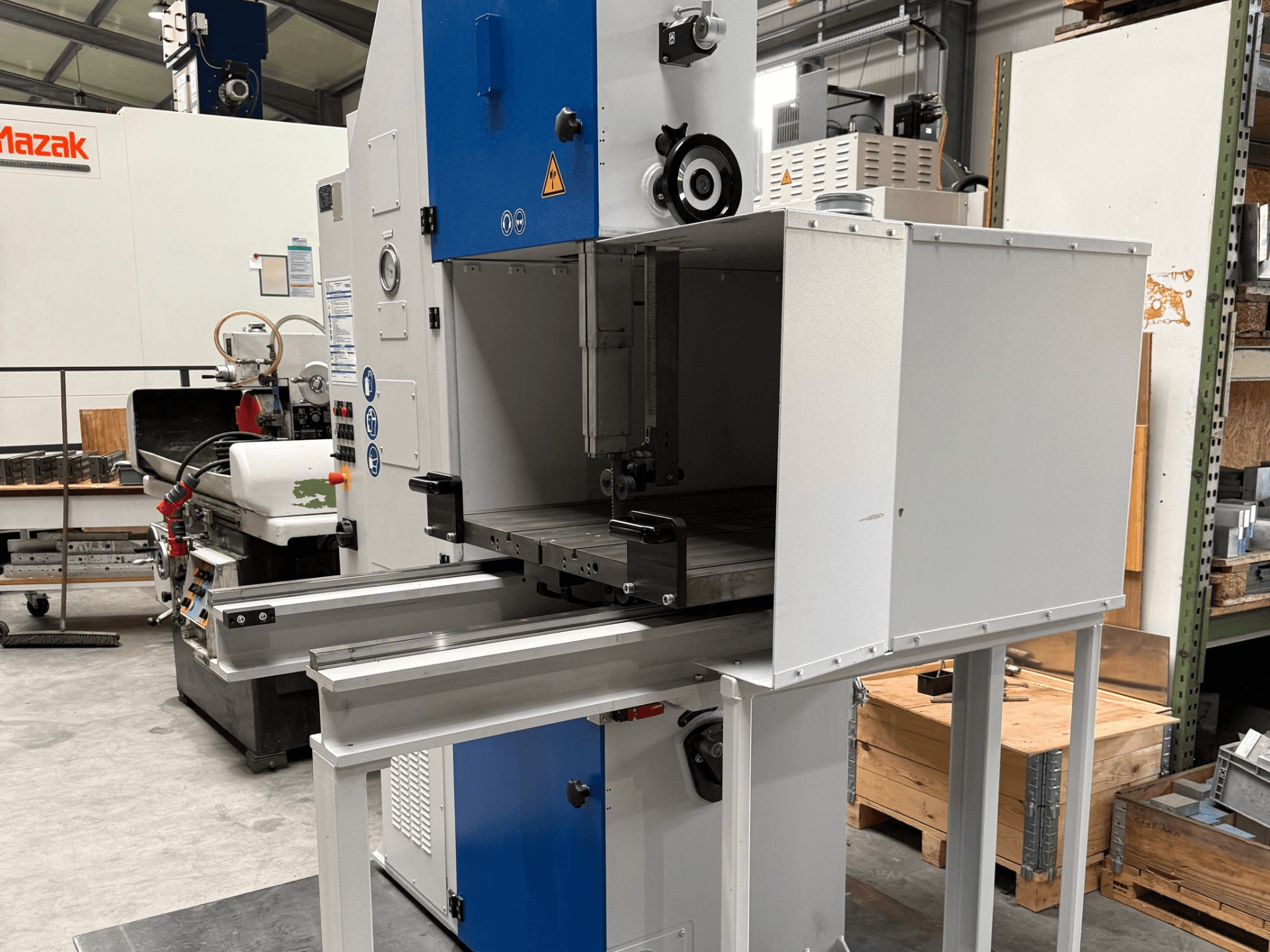 Máquina industrial Mazak vista de frente, con panel de control azul, mesa metálica y advertencias de seguridad.