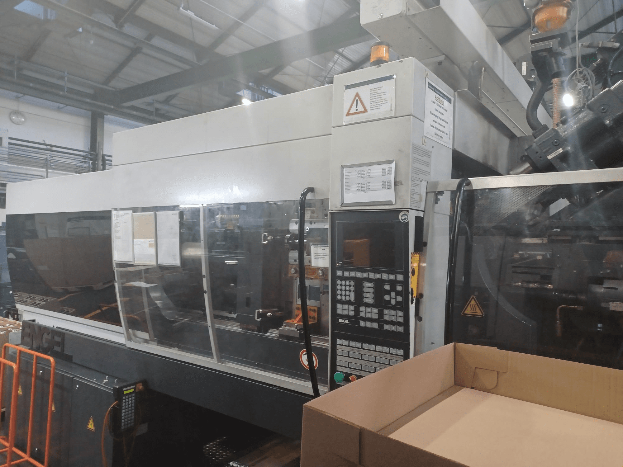 Máquina de moldeo por inyección Engel, vista frontal, con panel de control, etiquetas de seguridad y una caja vacía colocada cerca.