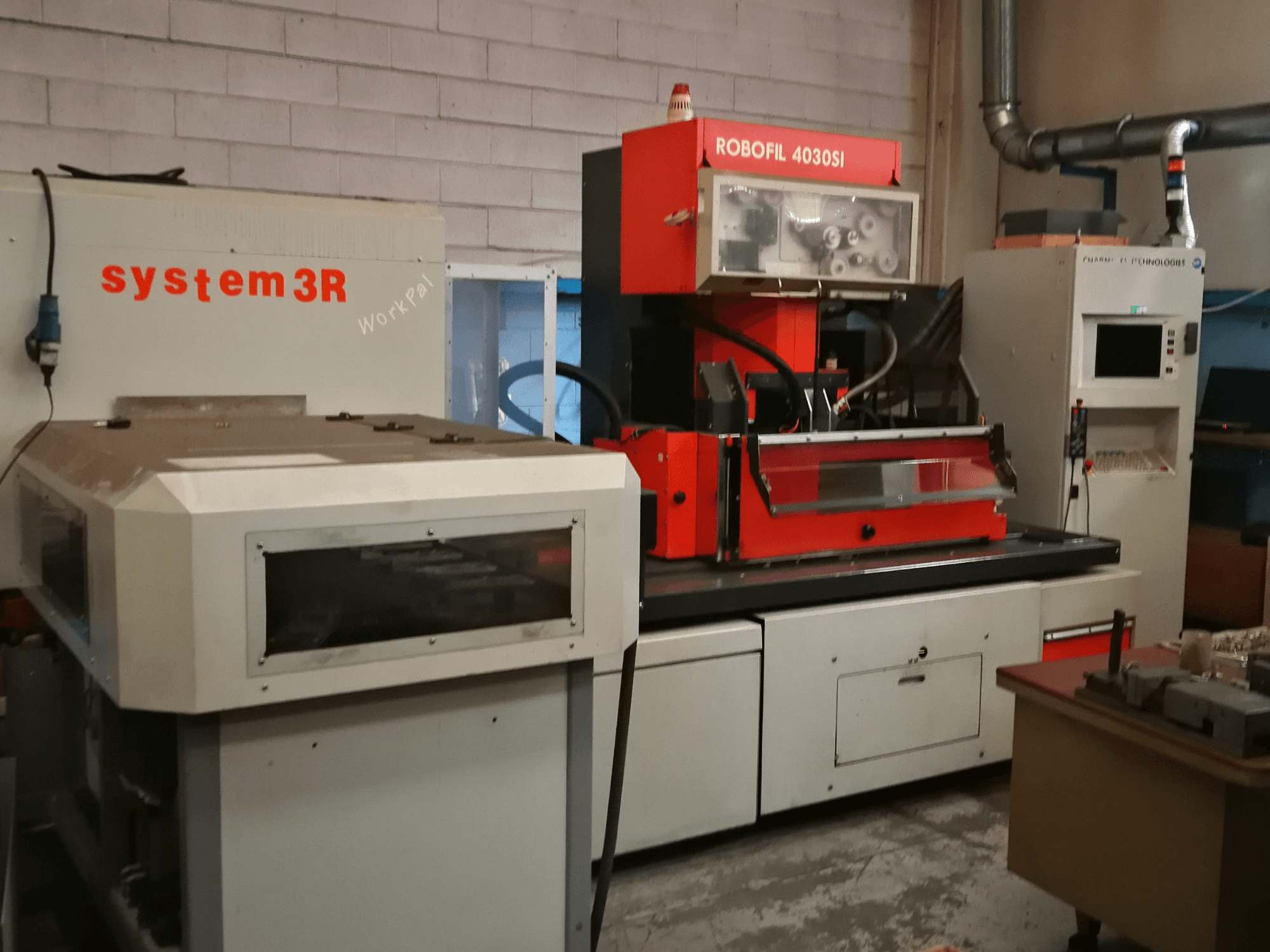 Vista frontal de la máquina de electroerosión por hilo ROBOFIL 4030 Si de CHARMILLES, con exterior rojo y gris y estación de trabajo system3R adyacente.