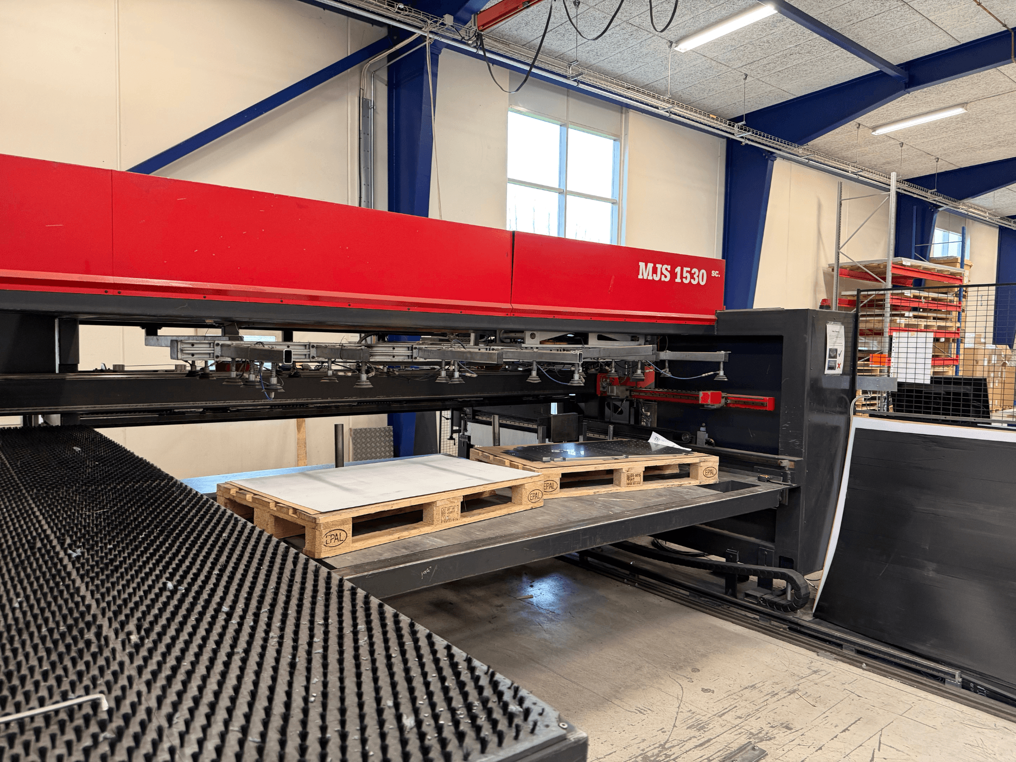 Máquina CNC MJS 1530 en un taller, vista frontal, con un palé de madera que sostiene una chapa metálica sobre una plataforma elevada.