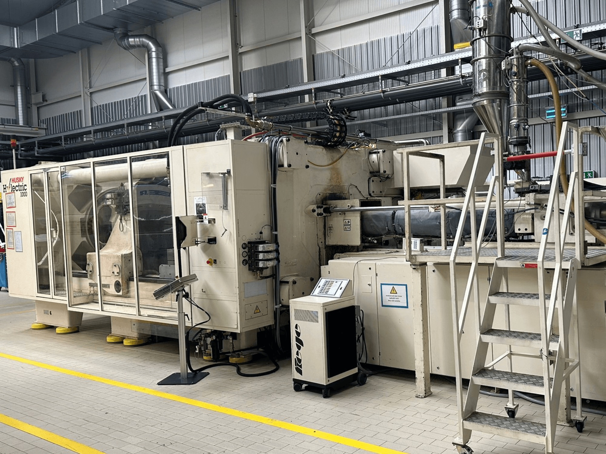 Vista frontal de la máquina de moldeo por inyección eléctrica Husky Hylelectric H1000 RS95/80 en un entorno industrial, en la que se aprecian el panel de control y los escalones.