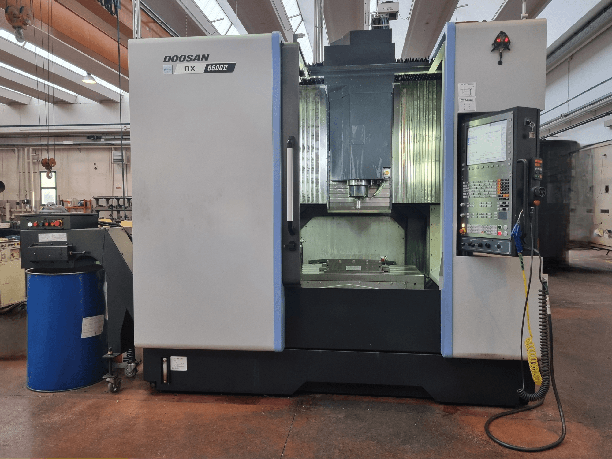 Máquina CNC Doosan NX 6500 II, vista frontal, con panel de control y zona de mecanizado, situada junto a un cañón azul.