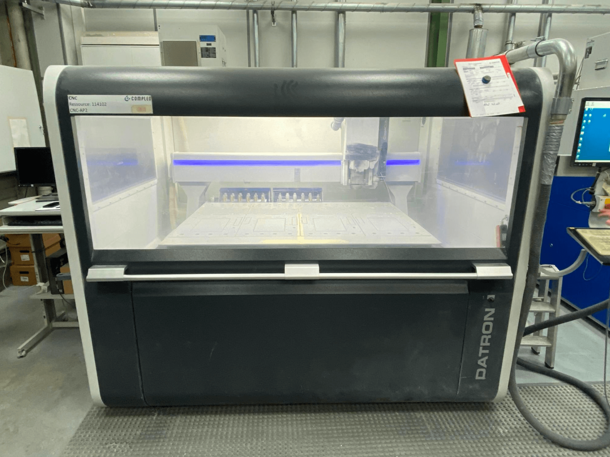 Vista frontal de un centro de mecanizado vertical Datron MLCube B, con controles visibles y una mesa de trabajo, en un entorno de taller.