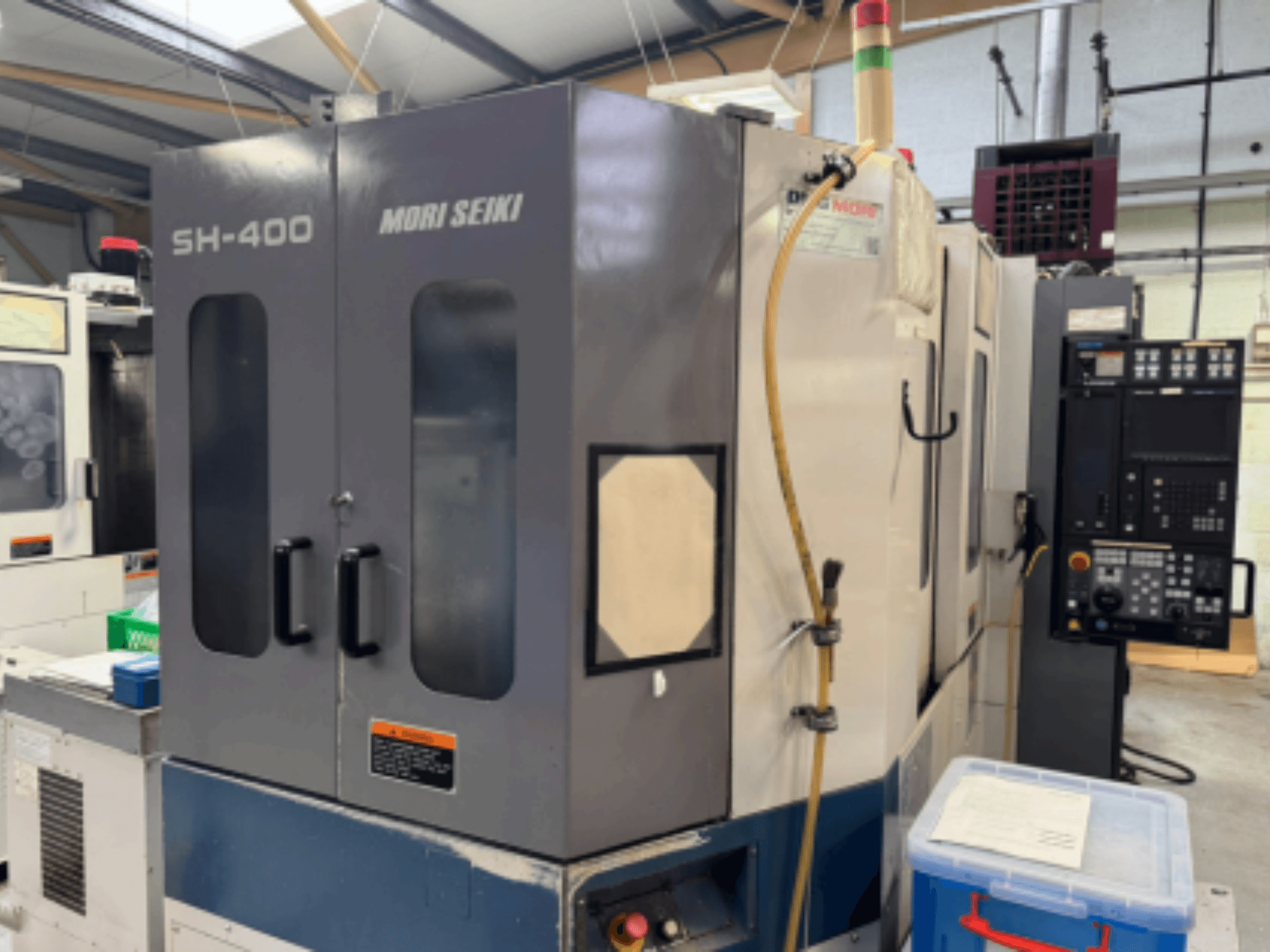 Máquina CNC Mori Seiki SH-400, vista frontal, con puertas dobles, panel de control y manguera de fijación. Contenedor de almacenamiento azul cerca.