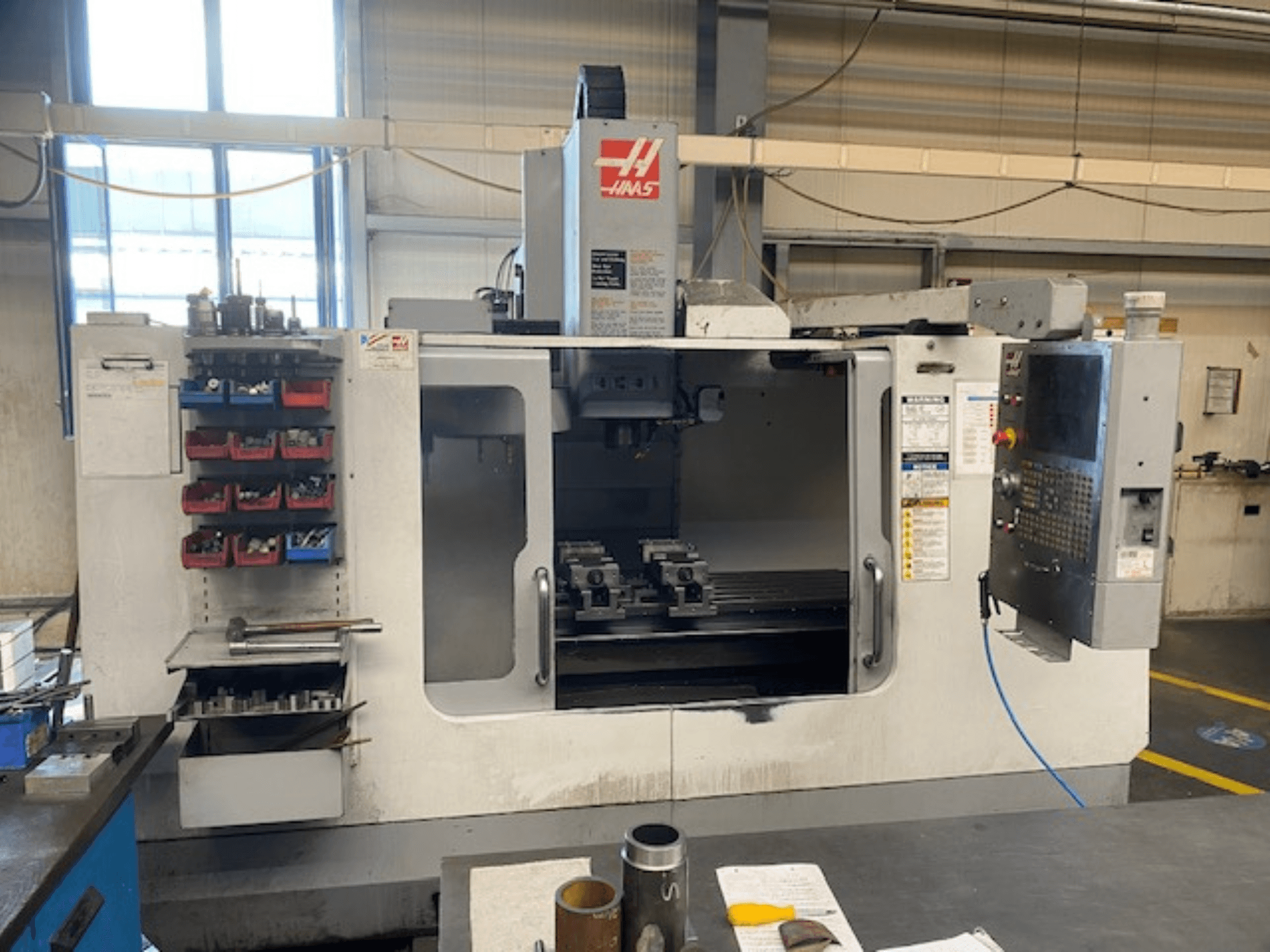 Vista frontal de la fresadora CNC Haas, modelo VF-2, con almacenamiento de herramientas, panel de control y herramientas del espacio de trabajo visibles.