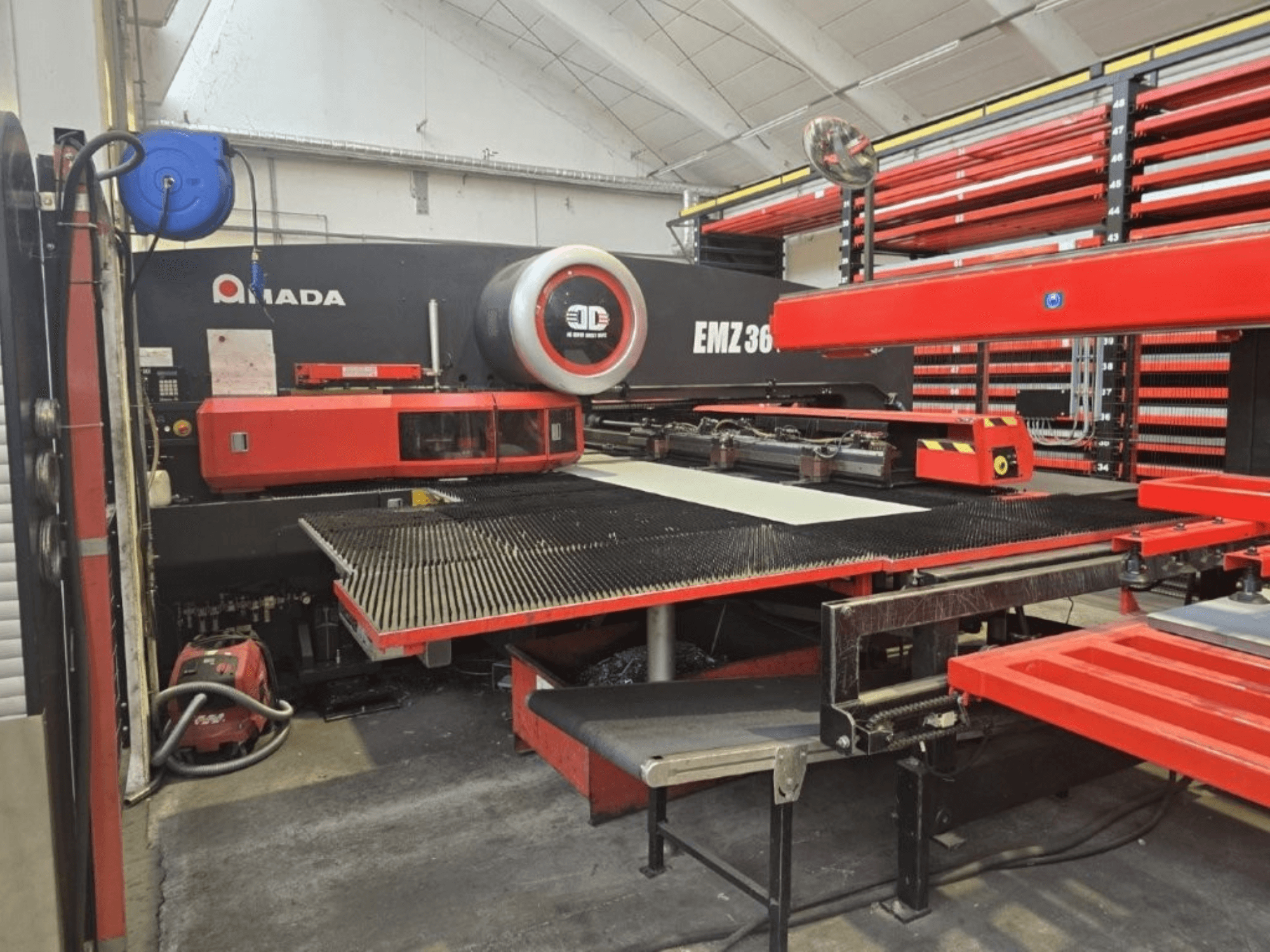 Máquina de corte por láser Amada EMZ 36 en un taller, vista frontal, con detalles en rojo y una gran mesa de corte.