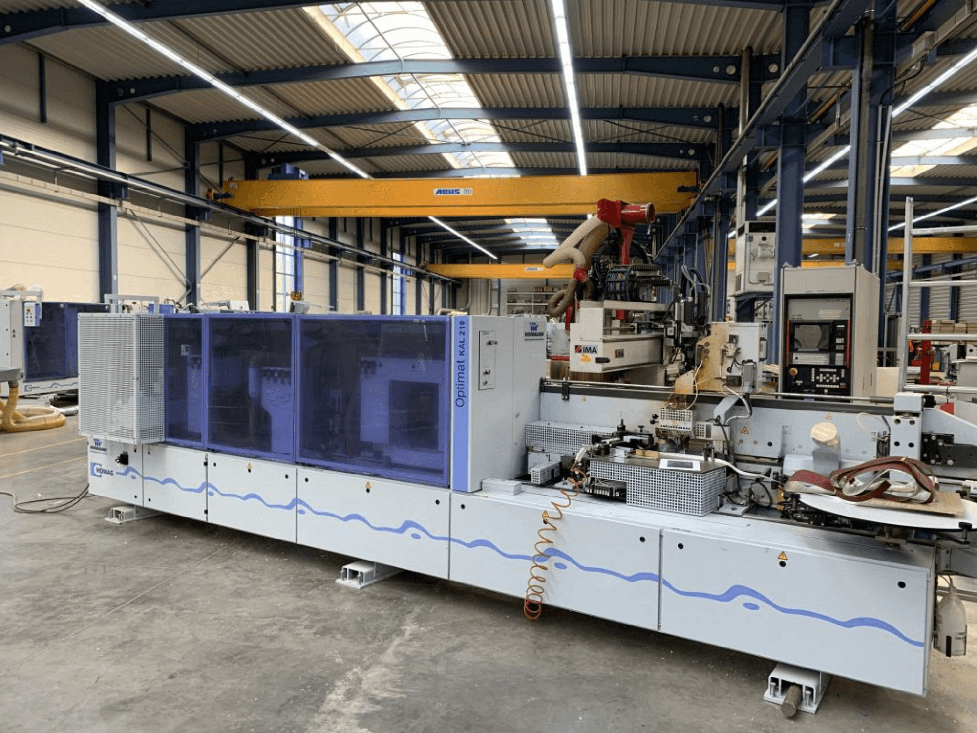 La encoladora de cantos HOMAG Optimat KAL210 vista desde delante, en la que se aprecian el panel de control y la zona de procesamiento en un entorno industrial.
