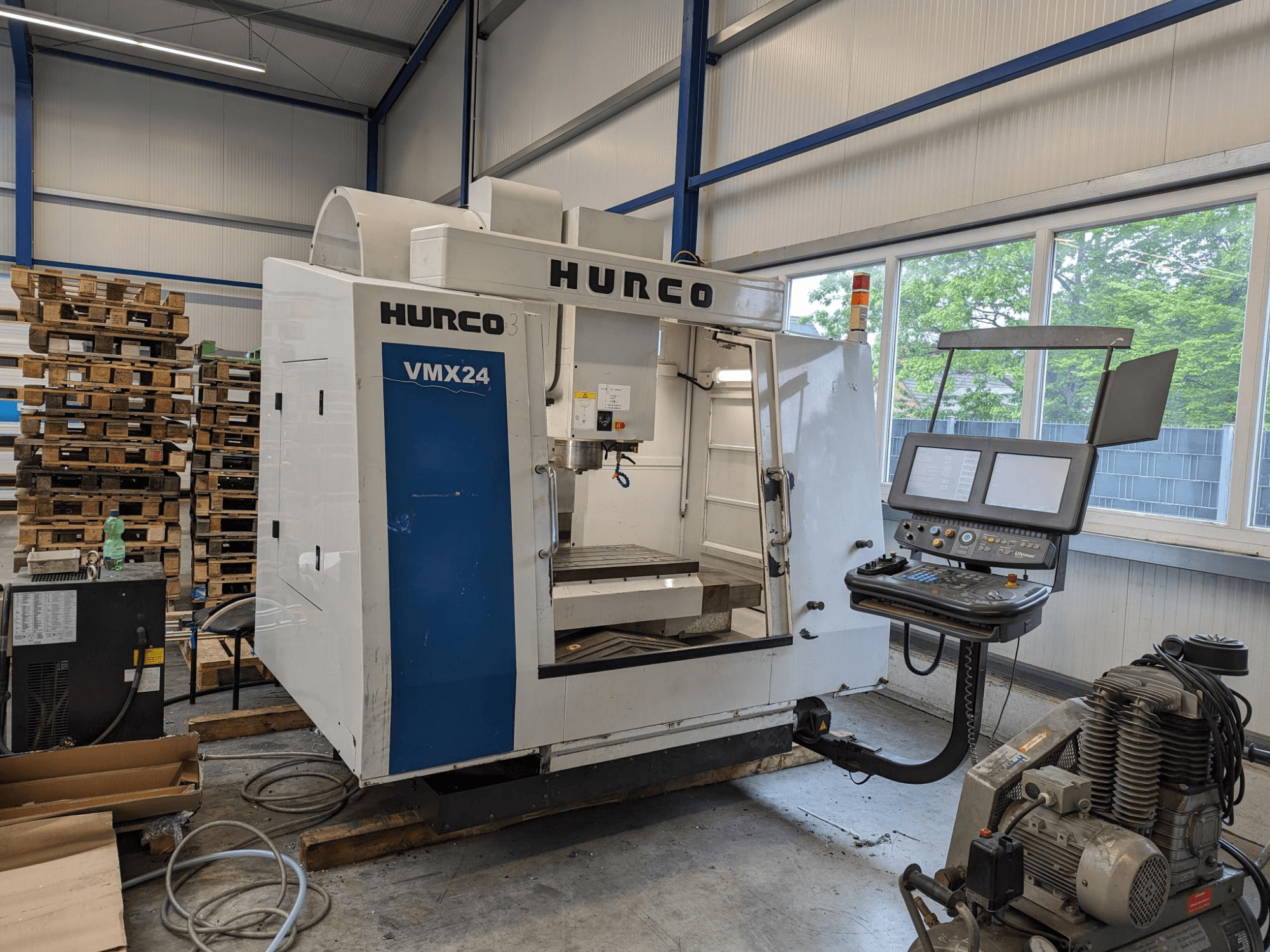 Fresadora Hurco VMX24, vista frontal, con panel de control y zona de herramientas; palets apilados al fondo; entorno de trabajo industrial.