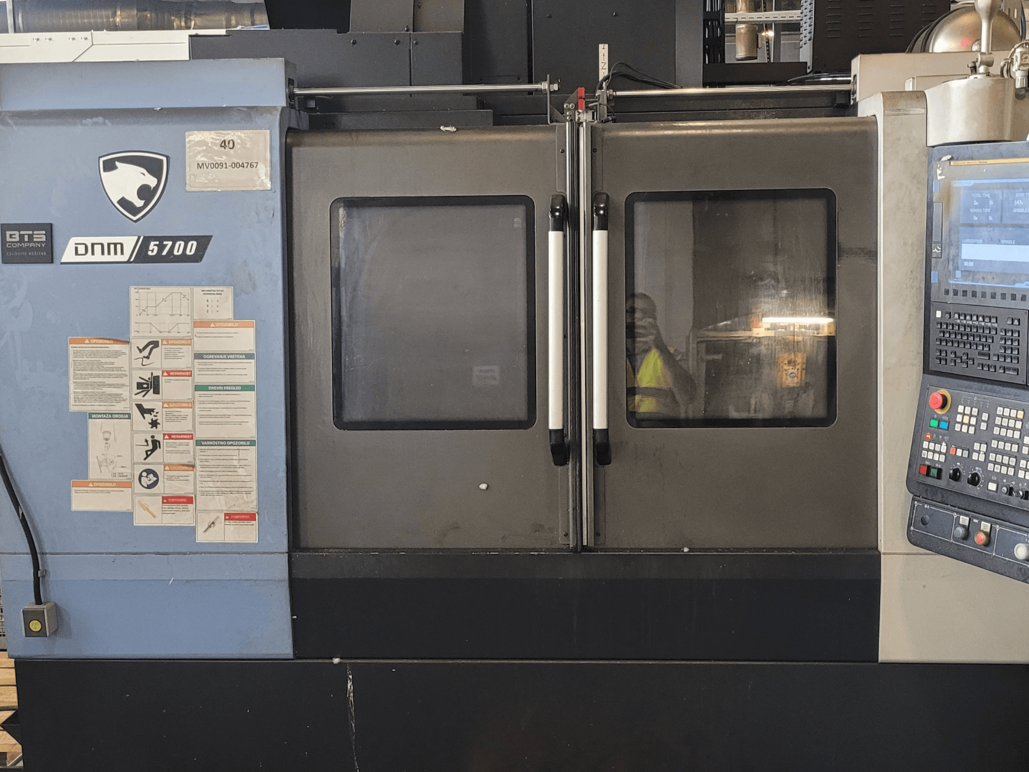 Vista frontal del centro de mecanizado vertical DOOSAN DNM 5700 con el panel de control, las etiquetas de seguridad y las puertas ligeramente entreabiertas.