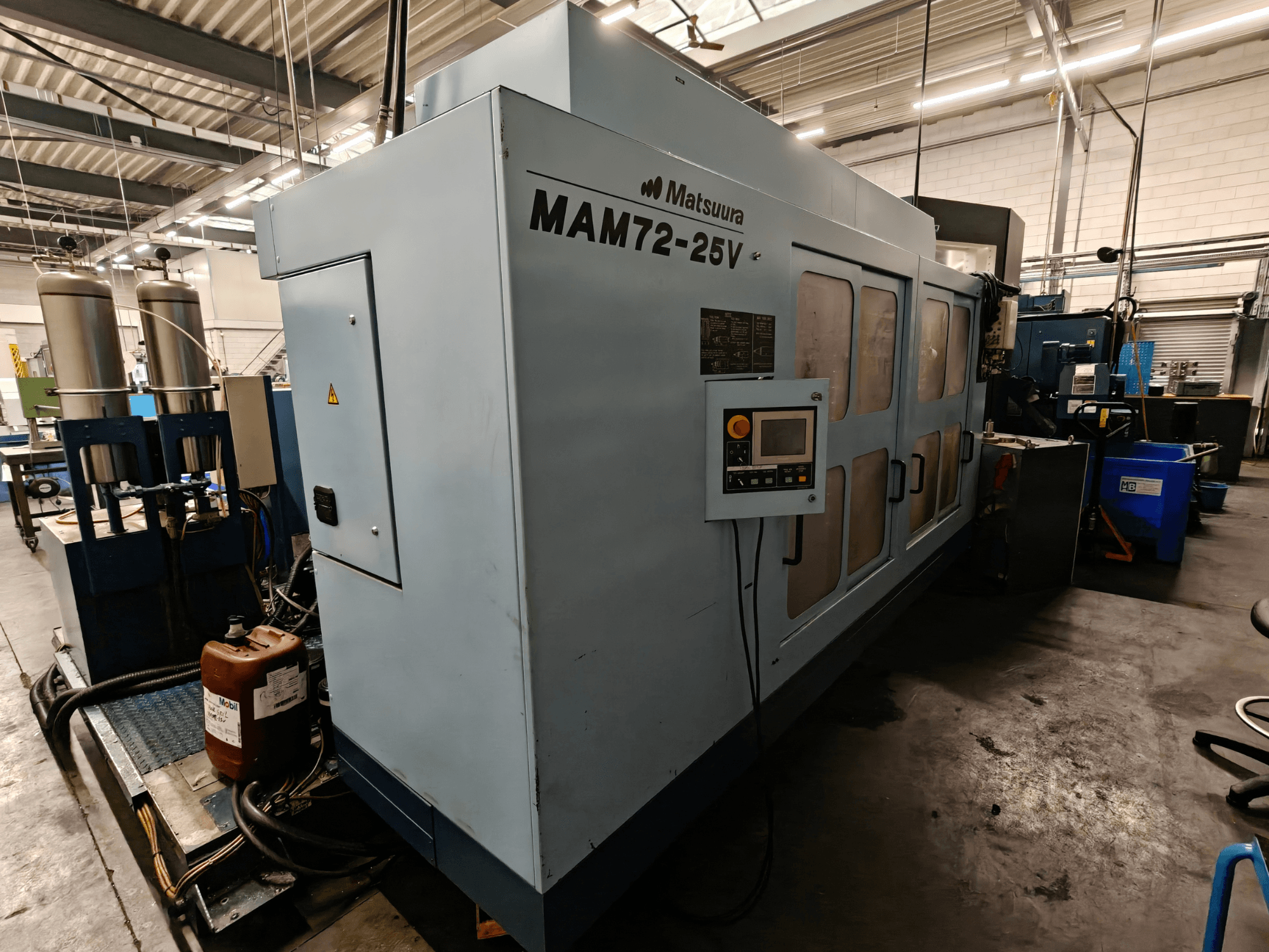 Máquina CNC Matsuura MAM72-25V, vista lateral, con panel de control, depósitos dobles de refrigerante y espacio de trabajo en un entorno industrial.
