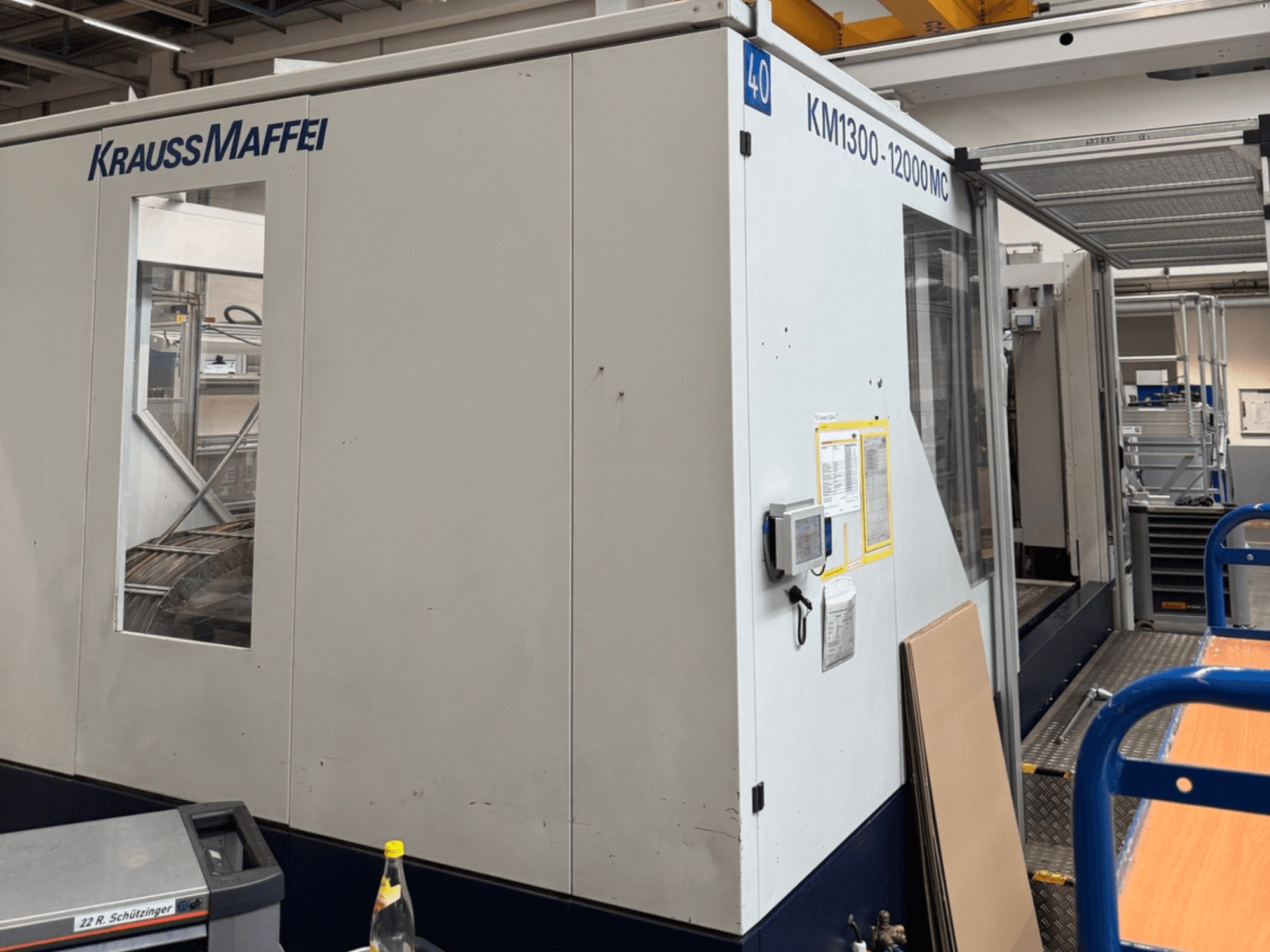 Máquina de moldeo por inyección hidráulica Krauss Maffei KM1300-12000 MC, vista lateral, mostrando el panel de control y el espacio operativo.