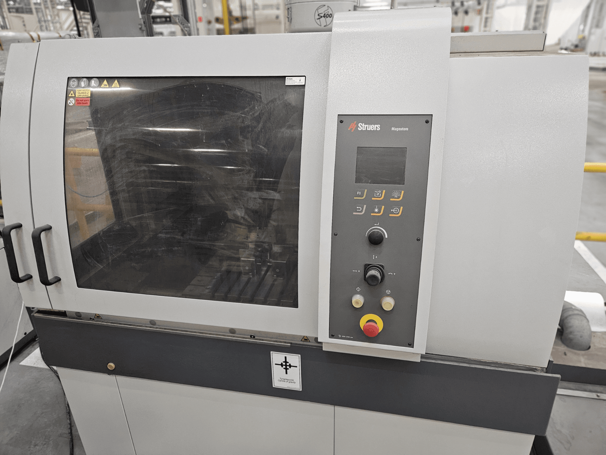 Vista frontal de la máquina Magpentor de Struers con panel de control, botones y una gran ventana de visualización. Etiquetas técnicas visibles.