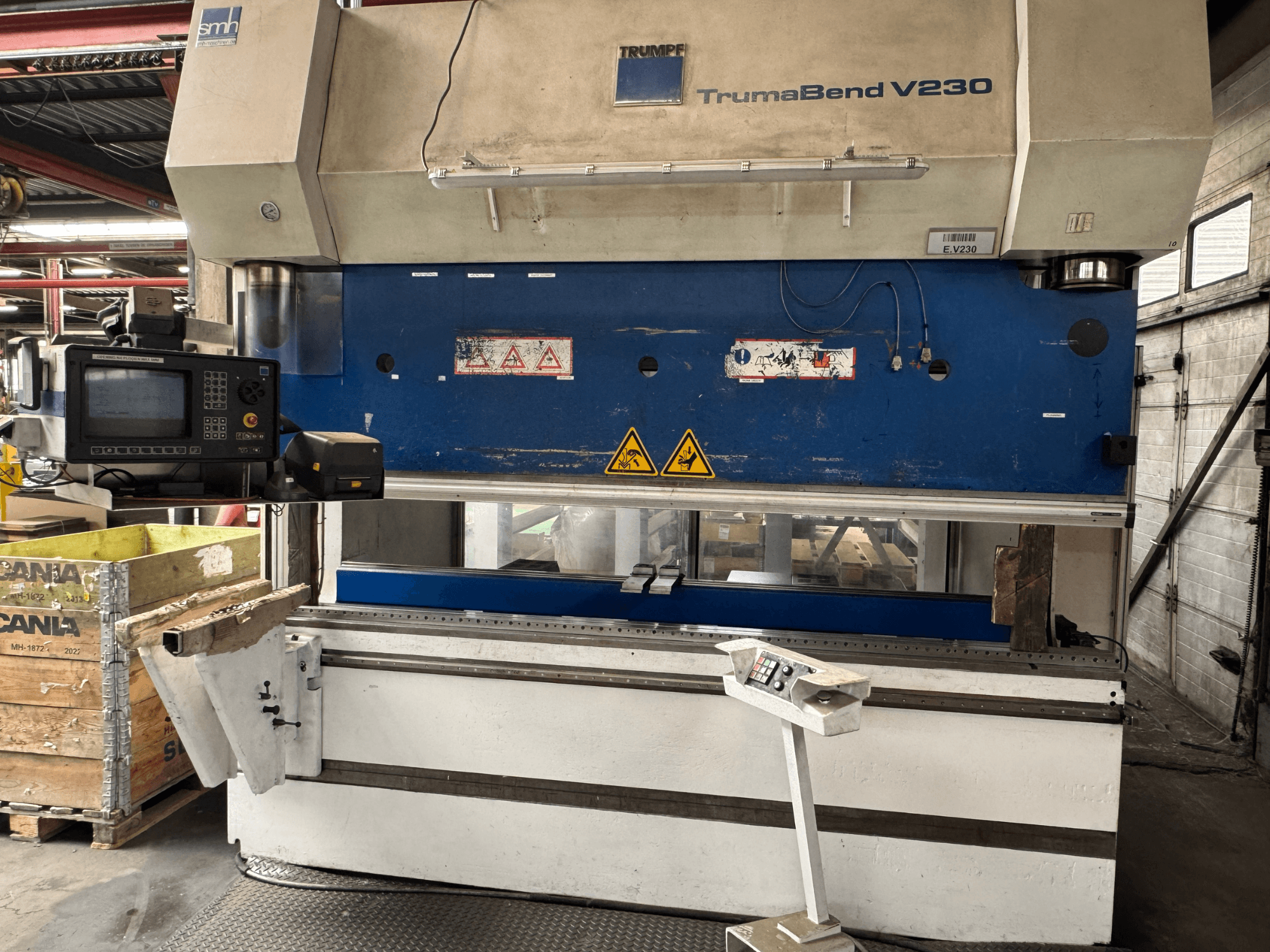 Plegadora TRUMPF TrumaBend V230, vista frontal con panel de control, etiquetas de advertencia y un brazo de soporte de la pieza de trabajo visibles.