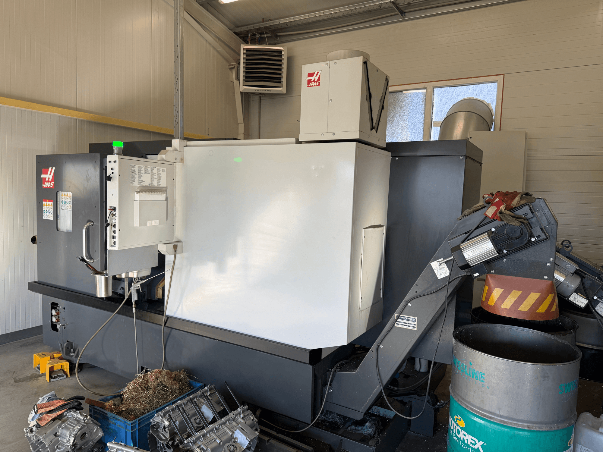 Máquina CNC Haas con panel de control, vista lateral; conductos de refrigerante y aceite visibles; desechos metálicos en un contenedor azul; entorno industrial.