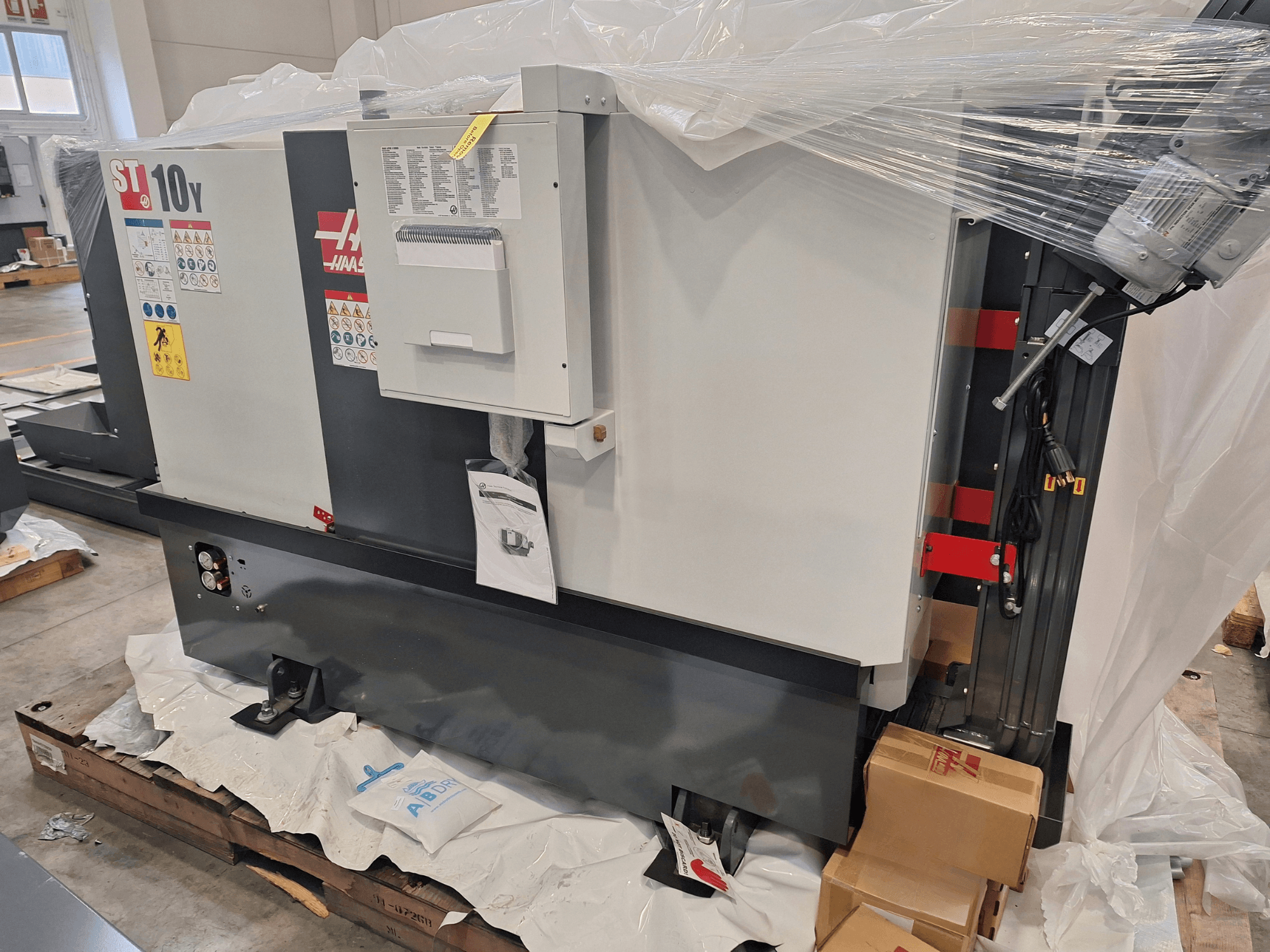 Vista lateral del torno horizontal HAAS ST-10Y EU, envuelto en plástico, con el panel de control y las etiquetas de advertencia visibles.