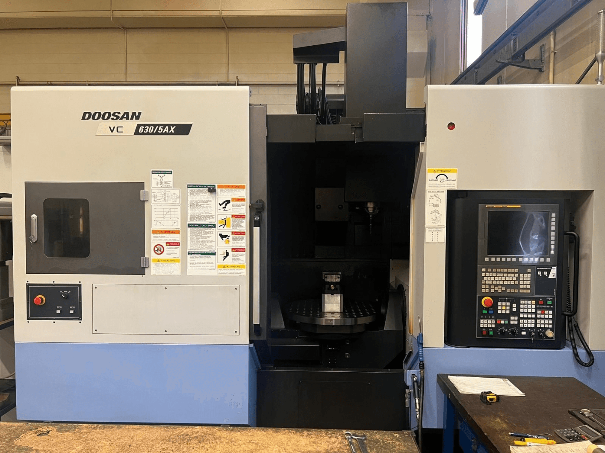 Vista frontal de un centro de mecanizado vertical DOOSAN VC 630/5AX, en la que se aprecian el panel de control y la zona del husillo, con las etiquetas de seguridad visibles.
