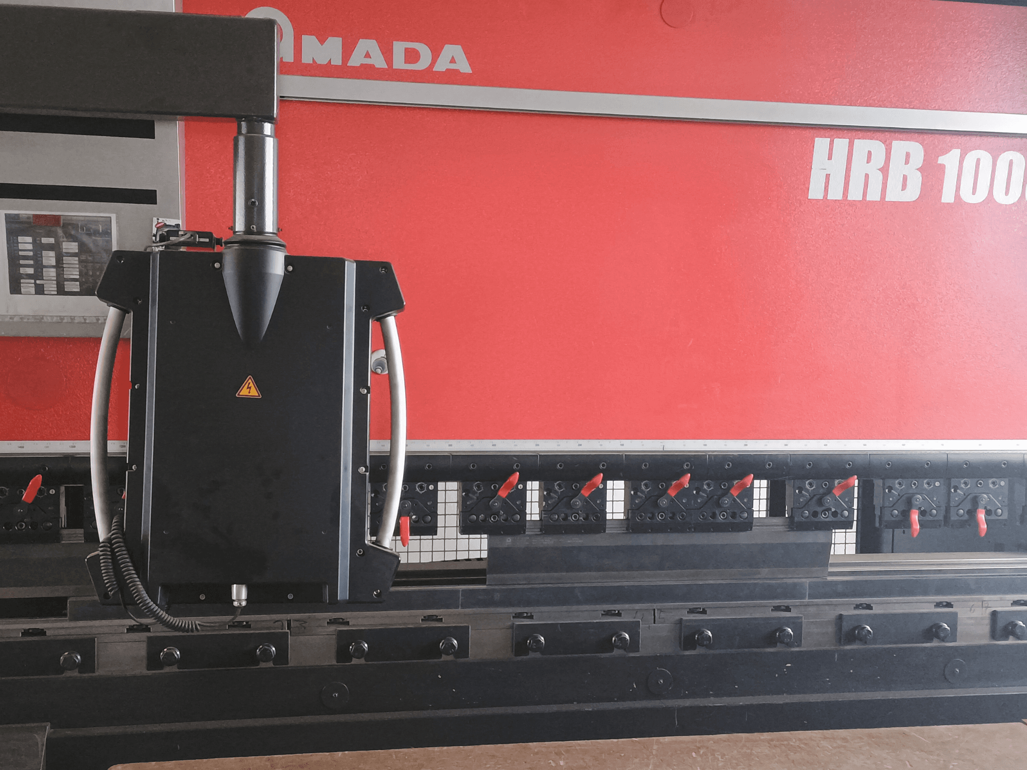 Vista frontal de la plegadora Amada HRB 100; incorpora un cabezal de control negro con una boquilla cónica y pinzas ajustables.