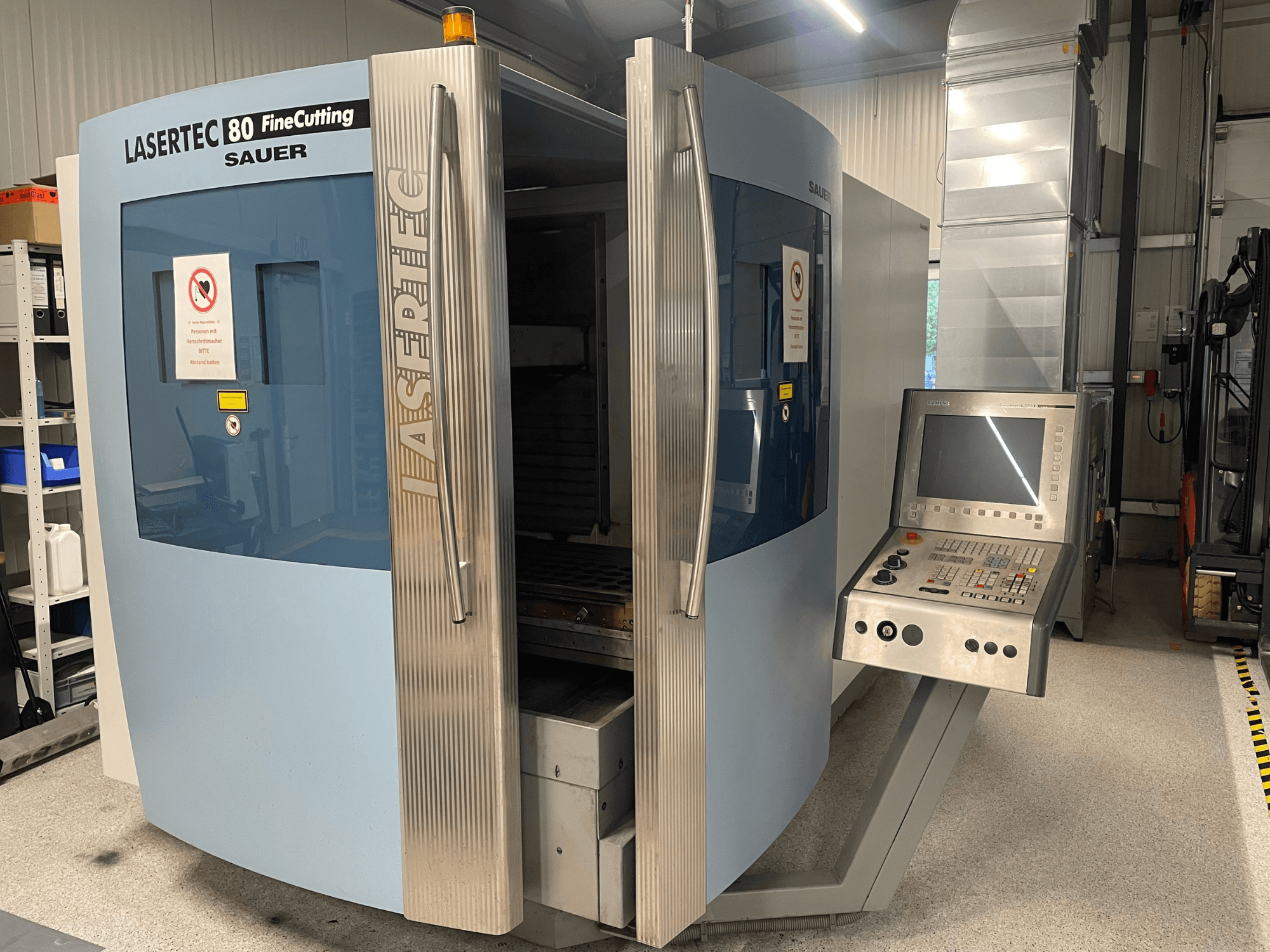 Vista frontal de la máquina para corte de chapa DMG Sauer Lasertec80 FineCutting, con el panel de control, las puertas abiertas y las señales de seguridad visibles.