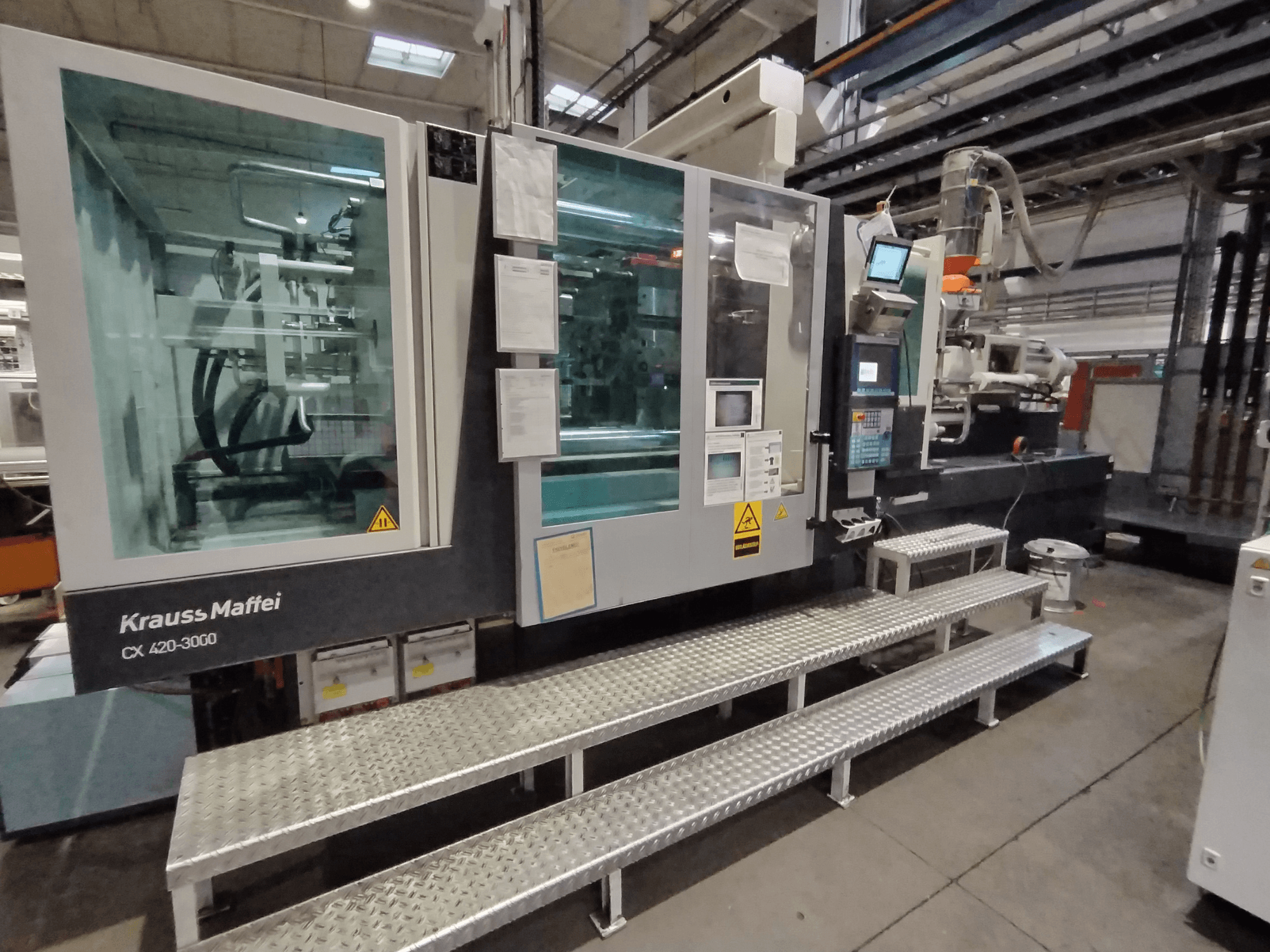 Máquina de moldeo por inyección Krauss Maffei CX 420-3000, vista lateral, con panel de control y señalización de seguridad visibles, colocada sobre escalones metálicos.