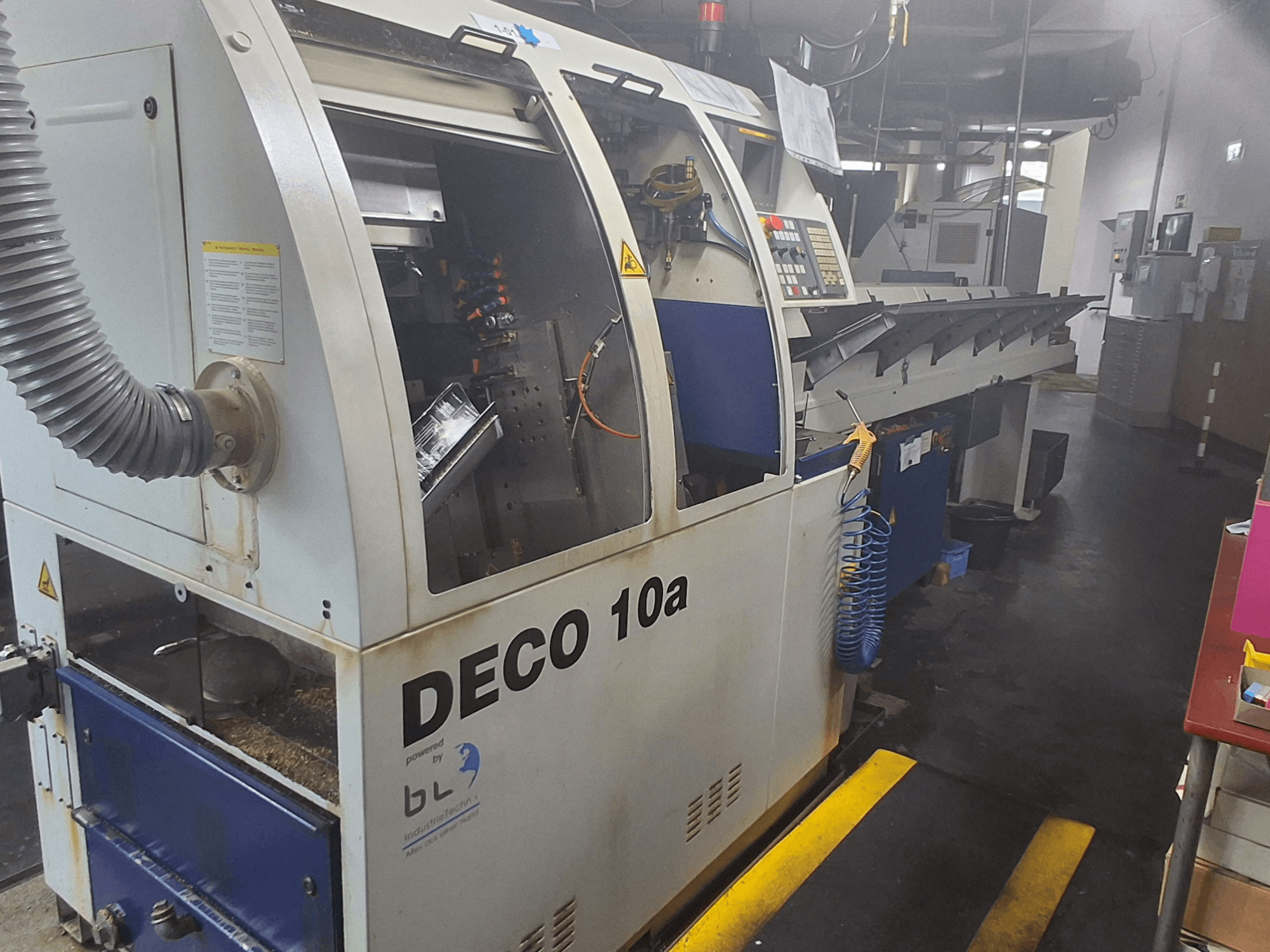 Máquina de torneado tipo suizo TORNOS DECO 10a, vista frontal; cuenta con un panel de control, mecanismos visibles y un tubo de extracción de polvo.