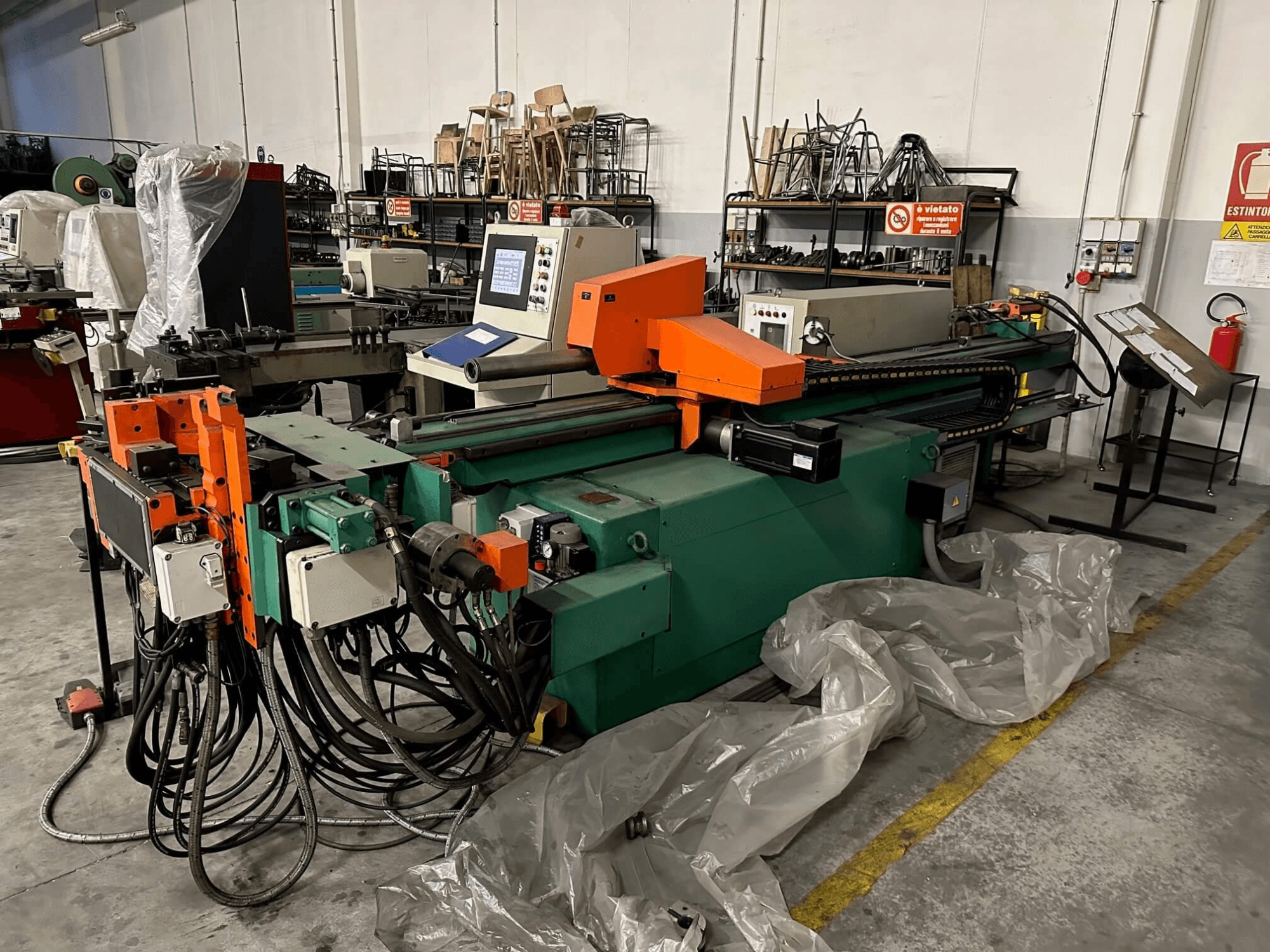 Máquina industrial con carrocería naranja y verde, panel de control y numerosos cables, vista de frente en un taller.