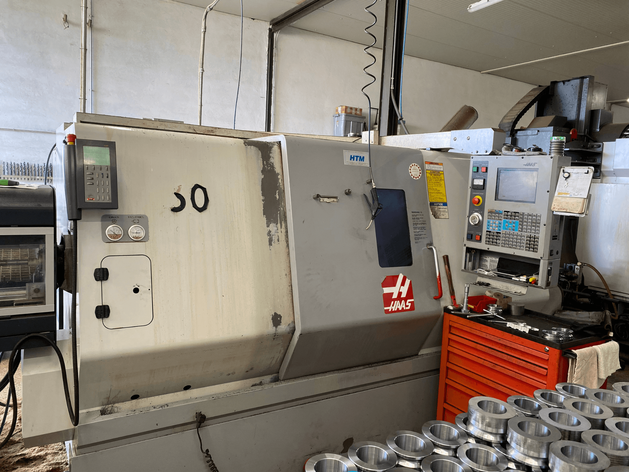 Máquina CNC Haas con panel de control, vista frontal, mostrando botones de operación, medidores y piezas metálicas apiladas cerca.