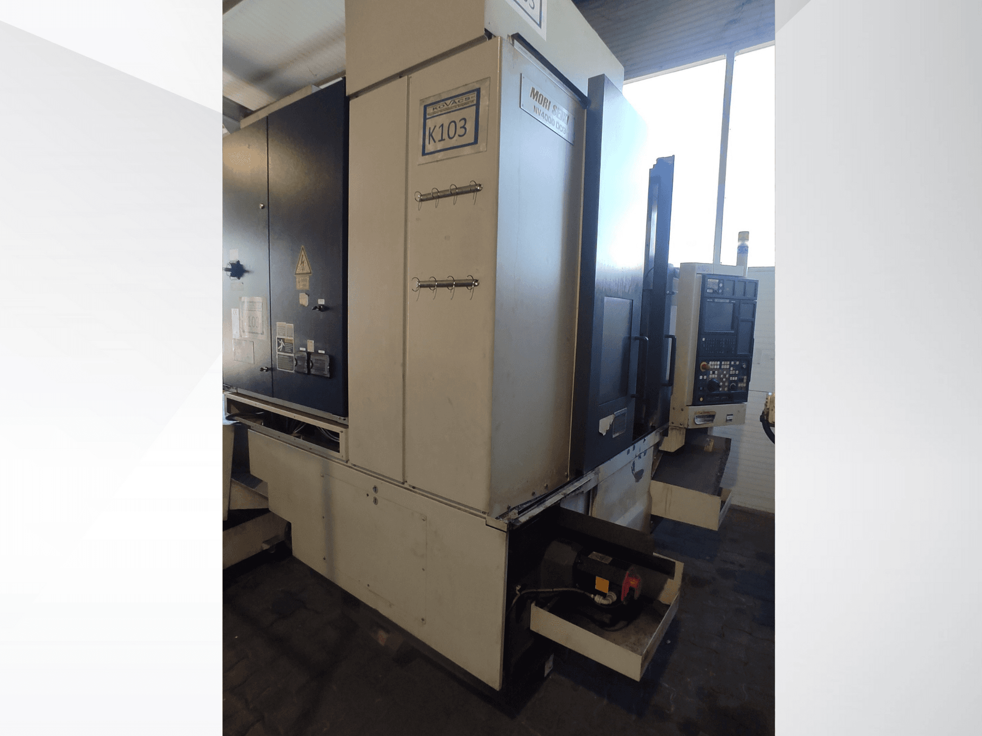 Máquina CNC Mori Seiki K103 en vista frontal, con panel de control, etiquetas de seguridad y componentes eléctricos visibles.