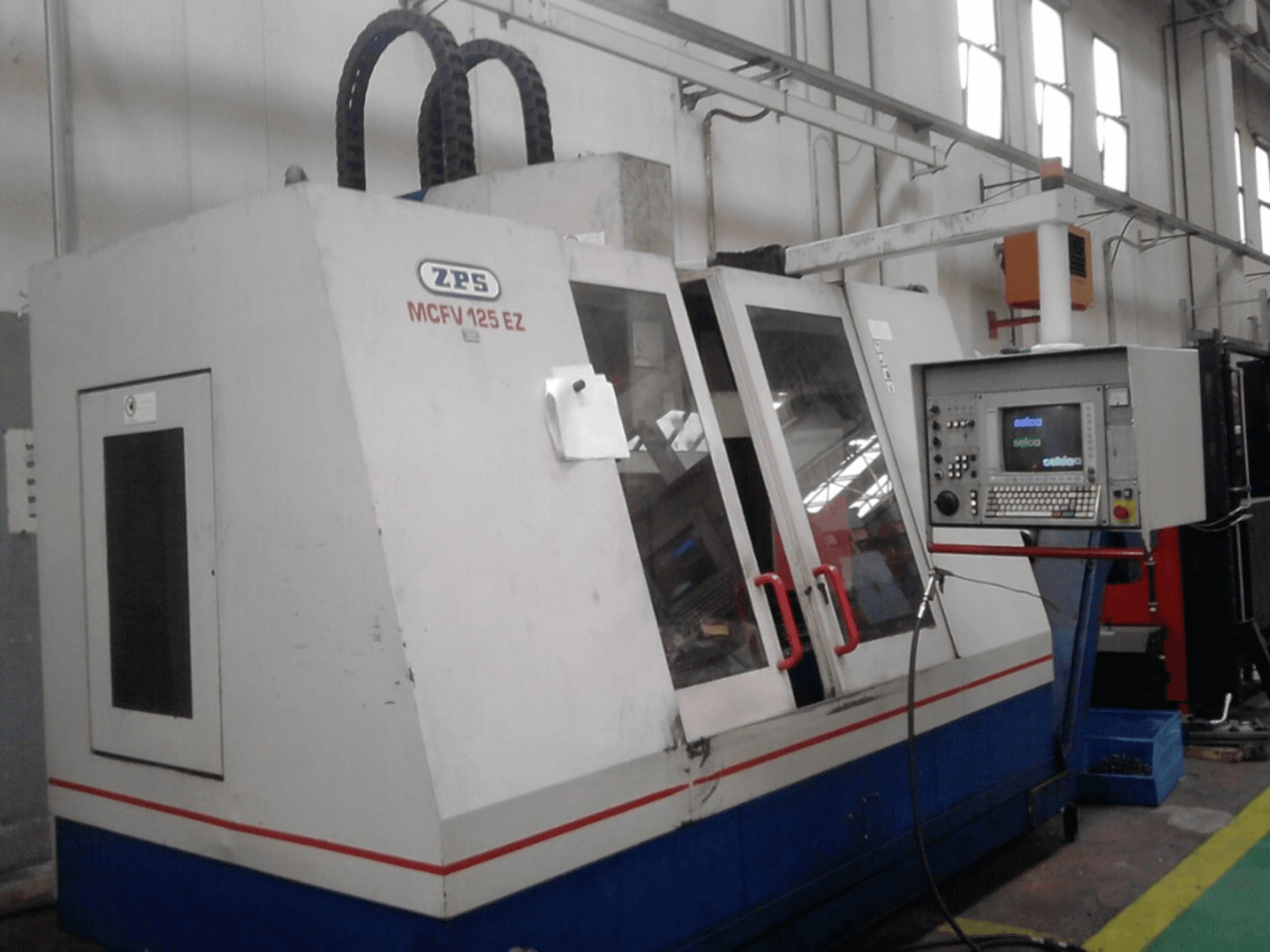 Máquina CNC ZPS MCFV 125 EZ, vista frontal con panel de control y asas rojas, en un taller con mangueras visibles.