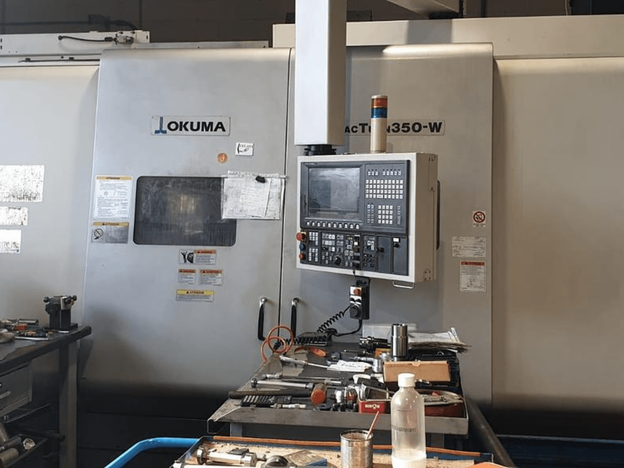 Centro de torneado y fresado Okuma MAC TURN 350-W, vista frontal con panel de control y espacio de trabajo abarrotado de herramientas y una botella.