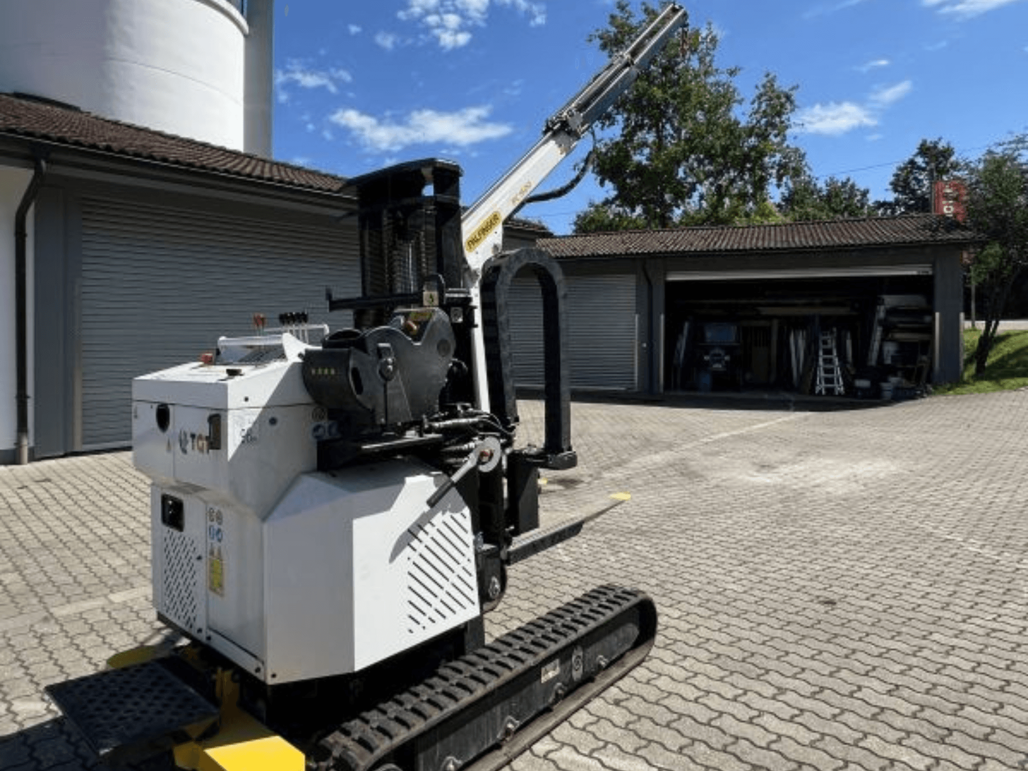 Minigrúa de construcción TGI, vista lateral, mostrando las orugas y la pluma montada, aparcada sobre una superficie de ladrillo junto a un garaje.
