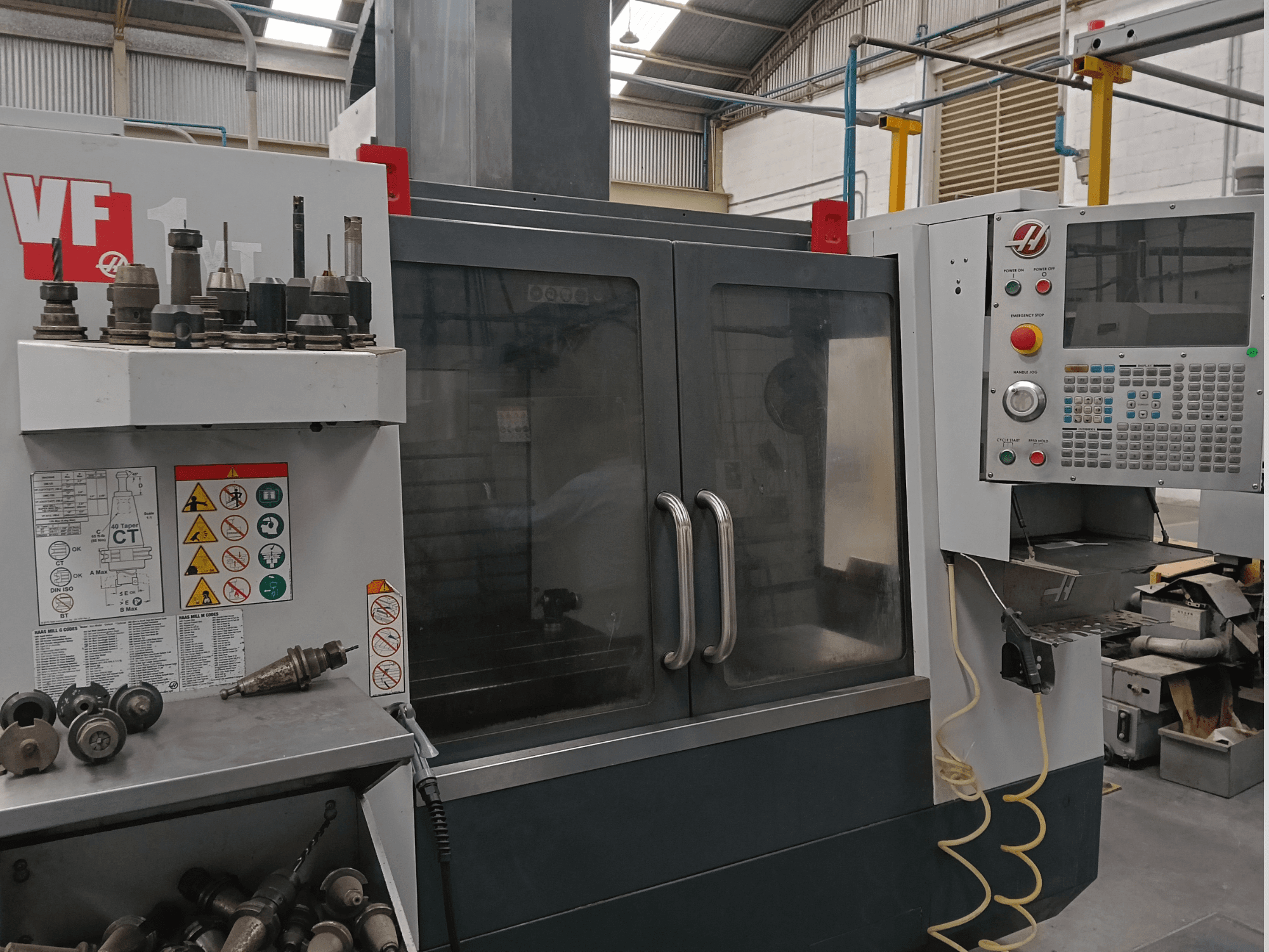 Centro de mecanizado vertical HAAS VF-1YT-3 en vista frontal, en el que se aprecian los mandos, los portaherramientas y las etiquetas de seguridad visibles.